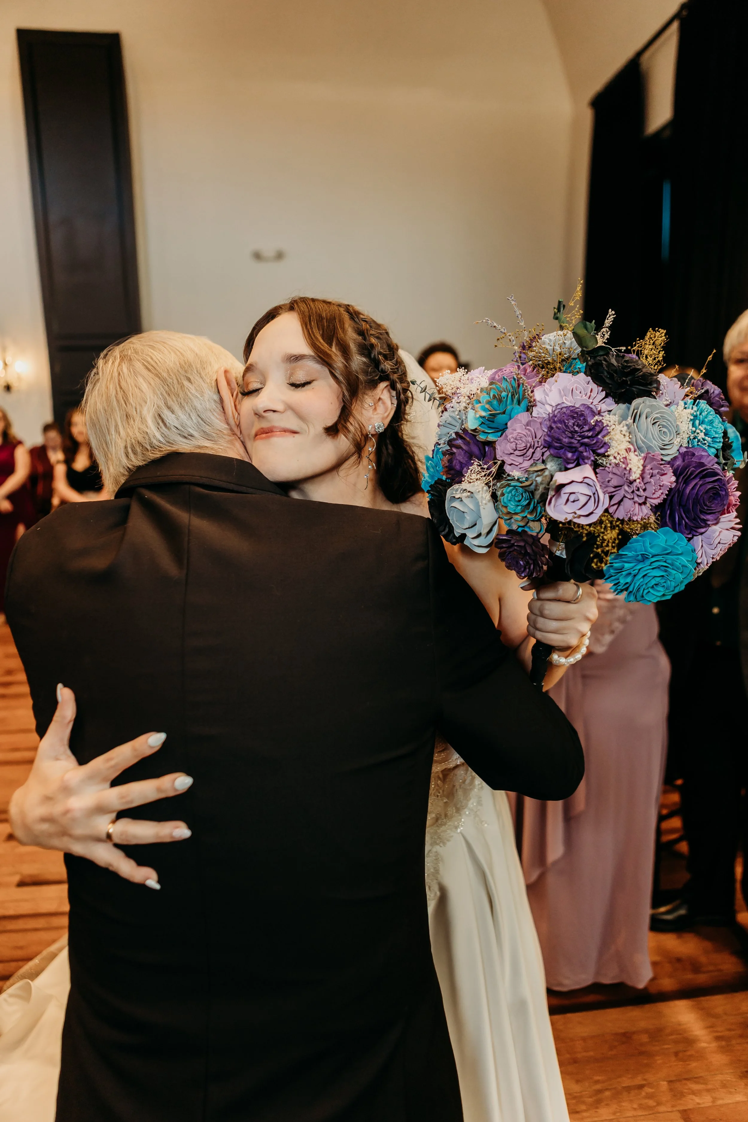 A bride hugging an older man, holding a bouquet of purple, blue, and lavender flowers, in a wedding reception setting.