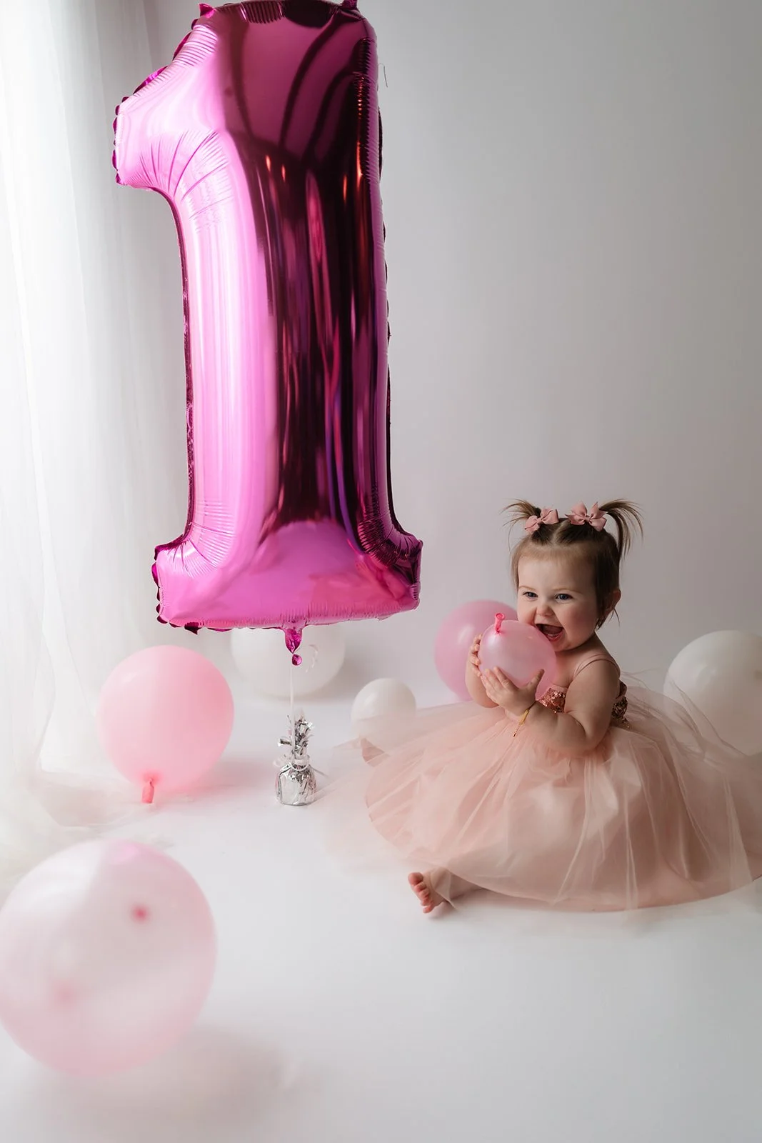 A toddler girl sitting on the floor in a pink tulle dress, holding a pink balloon, with pink and white balloons and a large pink number one foil balloon nearby, celebrating her first birthday.