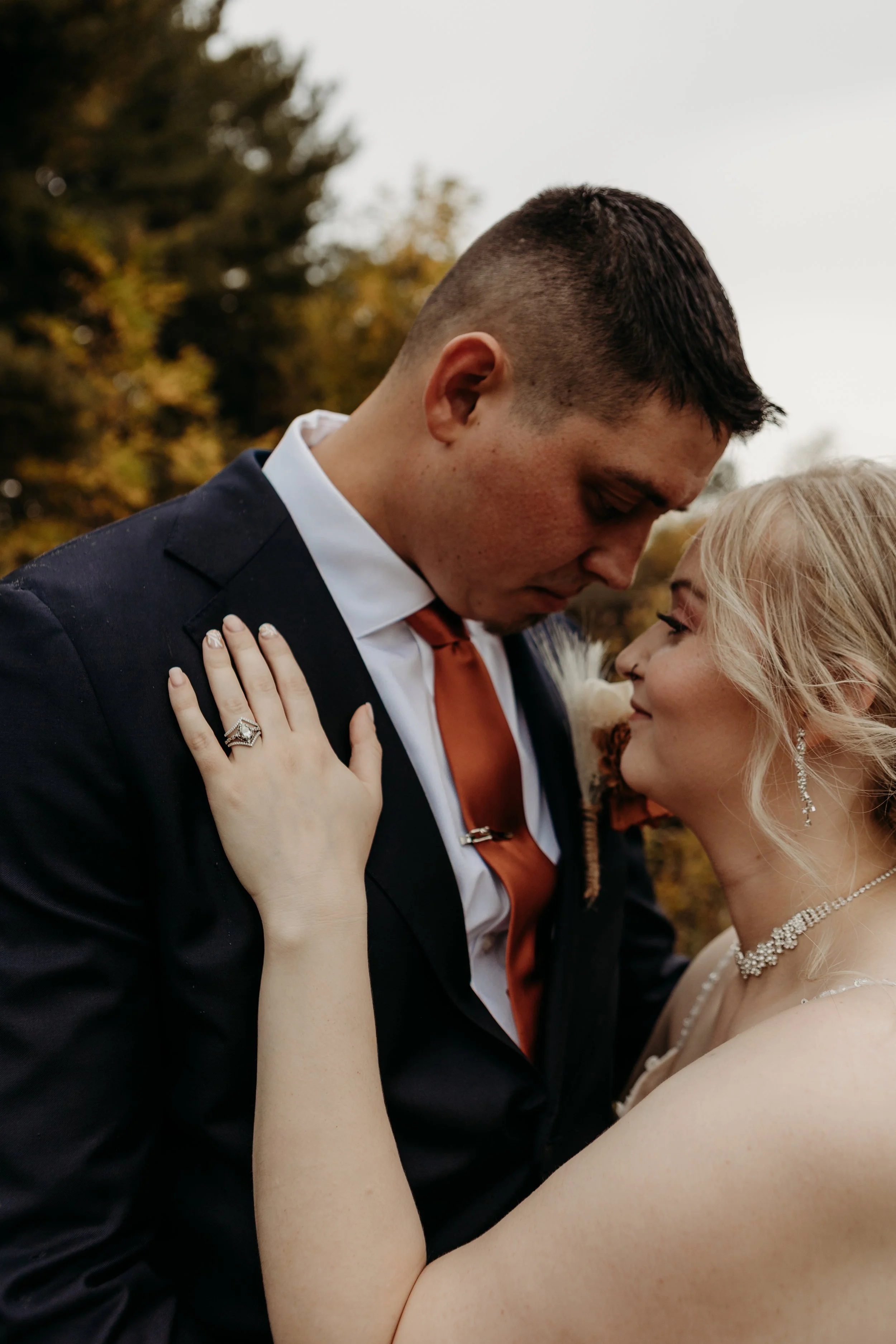 A bride and groom in wedding attire are close together, gazing into each other's eyes outdoors during autumn.