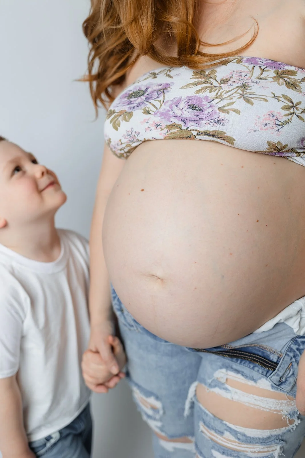 A pregnant woman with a floral top and ripped jeans holding hands with a young boy, both looking at each other.