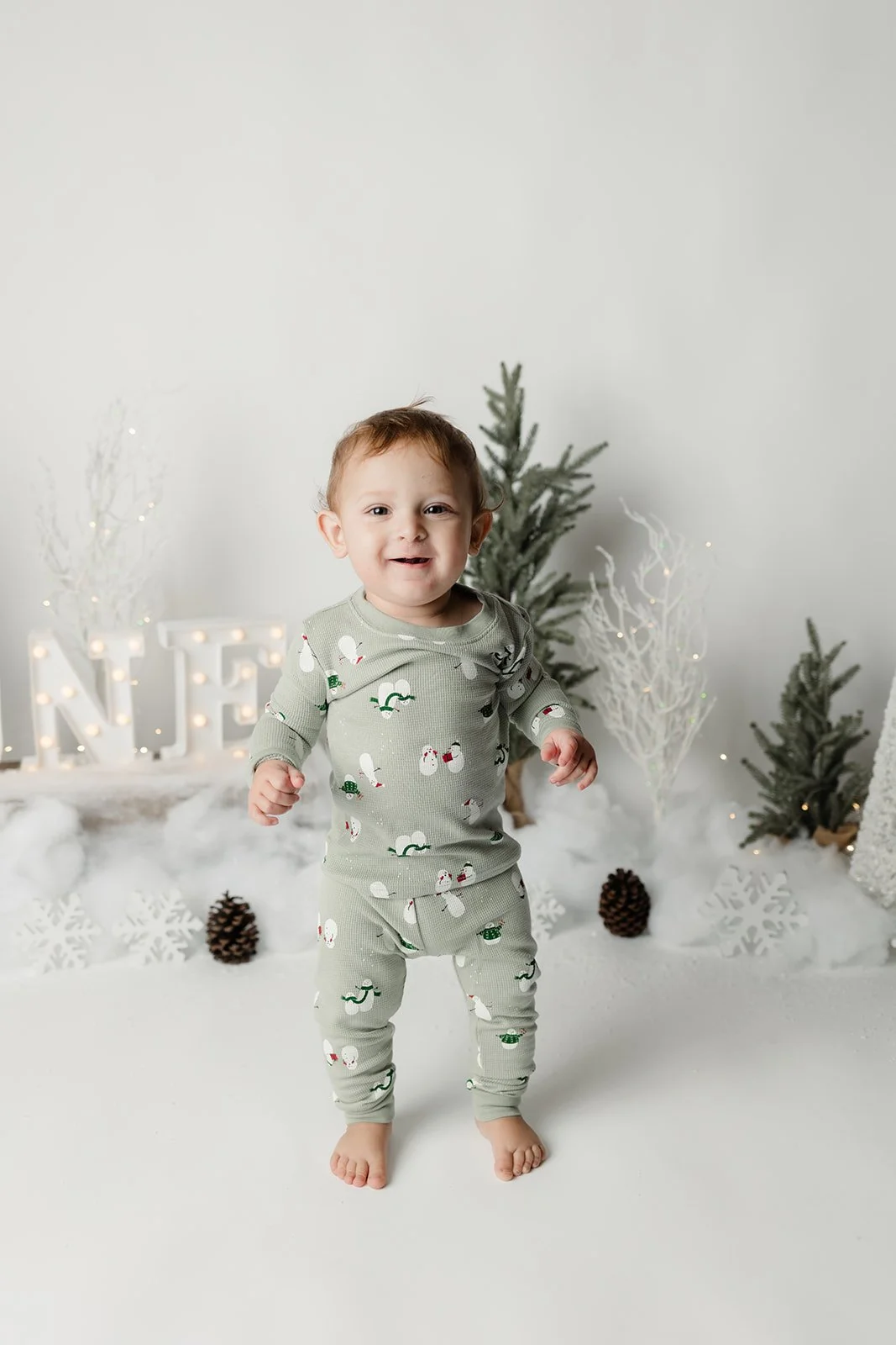 A young child with red hair standing in front of a winter holiday scene with small Christmas trees, pinecones, snow, and holiday decorations.