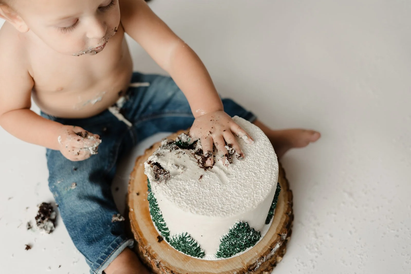 A young child with messy hands and face, wearing jeans, playing with a partially eaten birthday cake with white frosting and green decorations, sitting on a white surface.