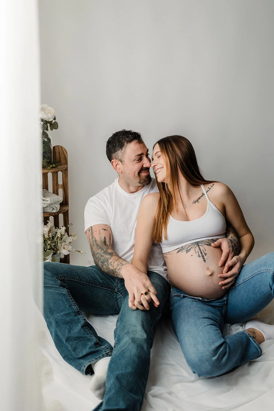 A couple sitting on a bed, facing each other smiling, with the woman showing her pregnant belly.