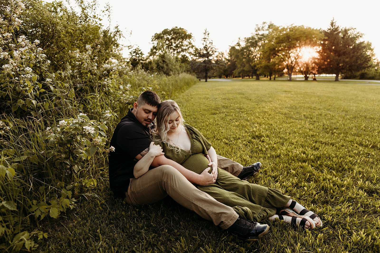 A couple sitting on the grass at sunset, with a pregnant woman and a man leaning against her, surrounded by greenery and trees in the background.