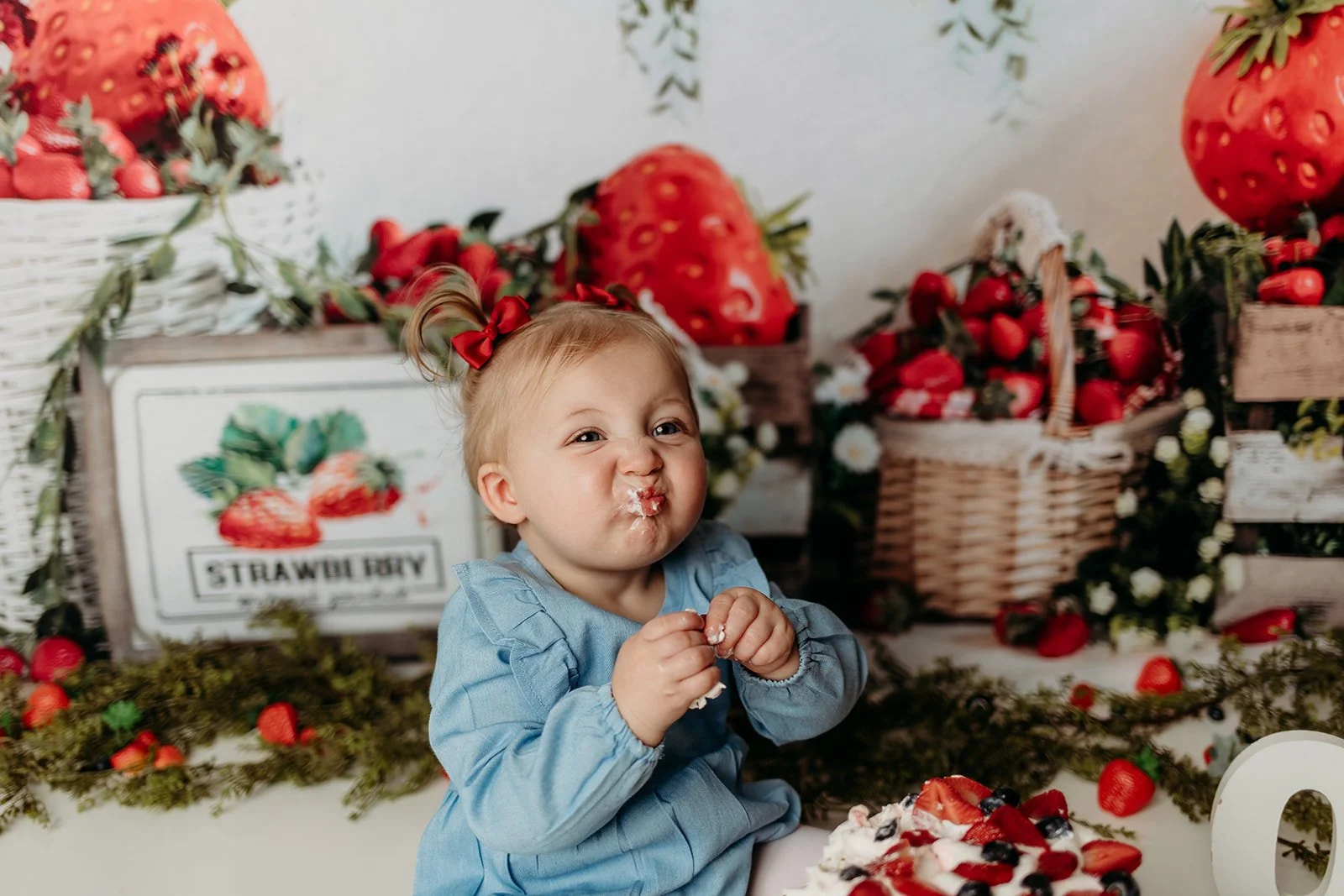 A young girl with blonde hair tied in a ponytail with a red bow, wearing a light blue dress, making a funny face with her cheeks puffed out and a bit of whipped cream on her face, sitting in front of a strawberry-themed backdrop with baskets of straw
