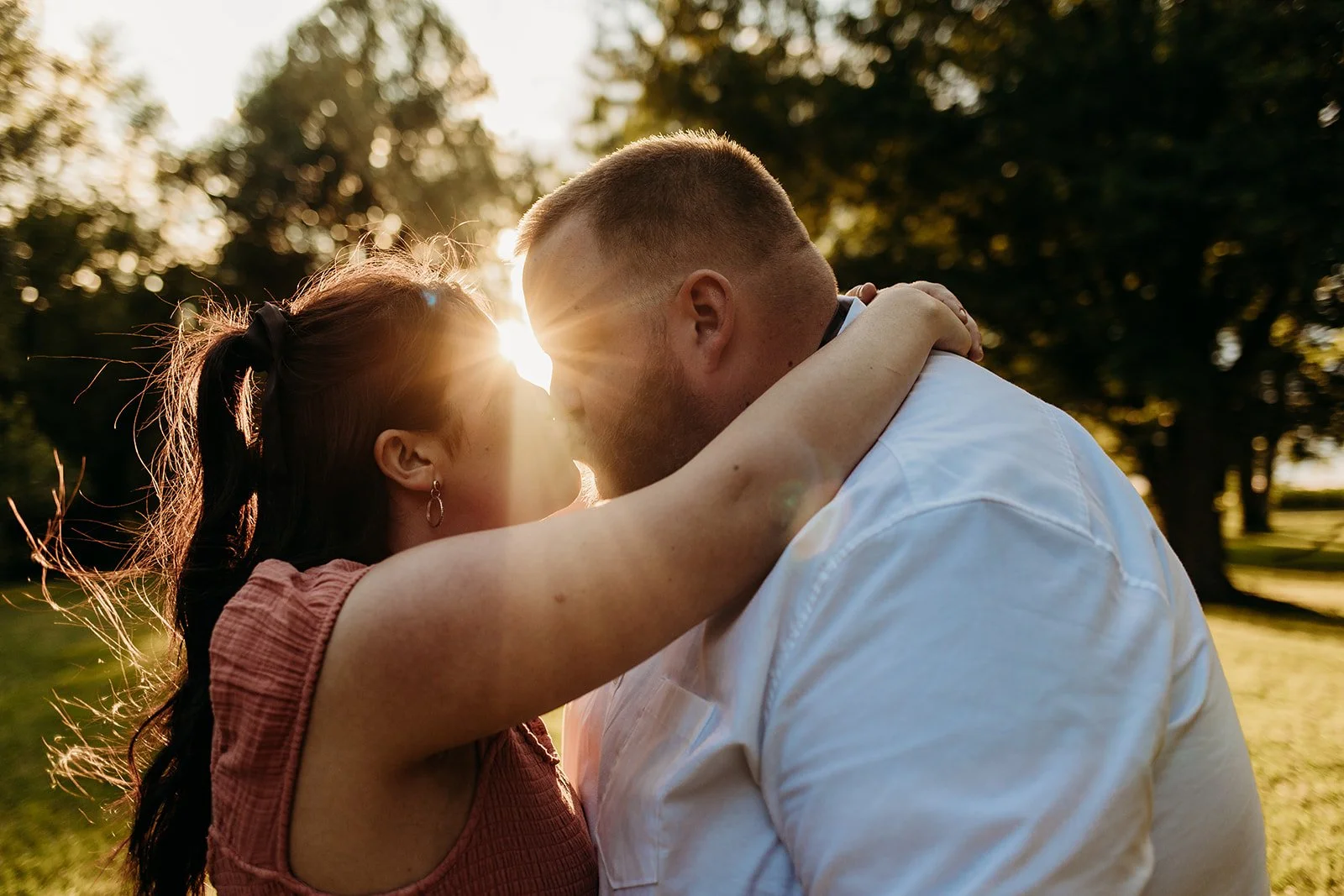 A couple embracing outdoors at sunset, with their foreheads touching and the sun shining between them.