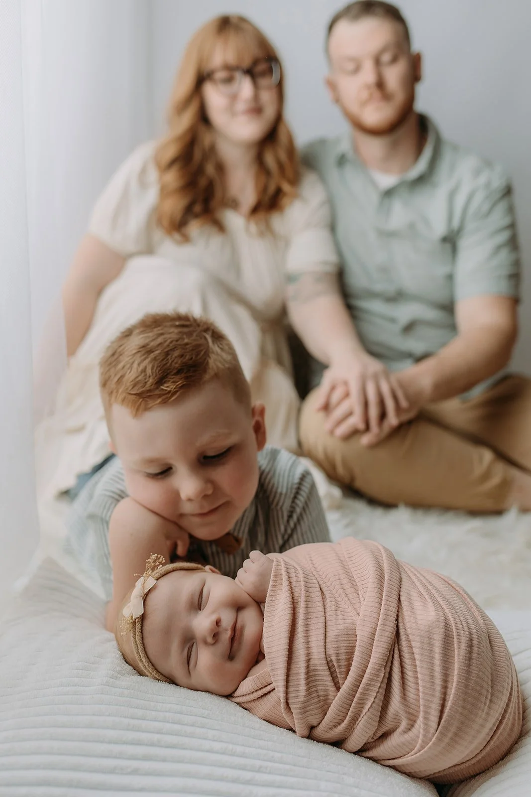 A family of four sitting on a bed, with a newborn baby in the foreground wrapped in a pink blanket, a young boy looking at the baby, and two adults in the background smiling.