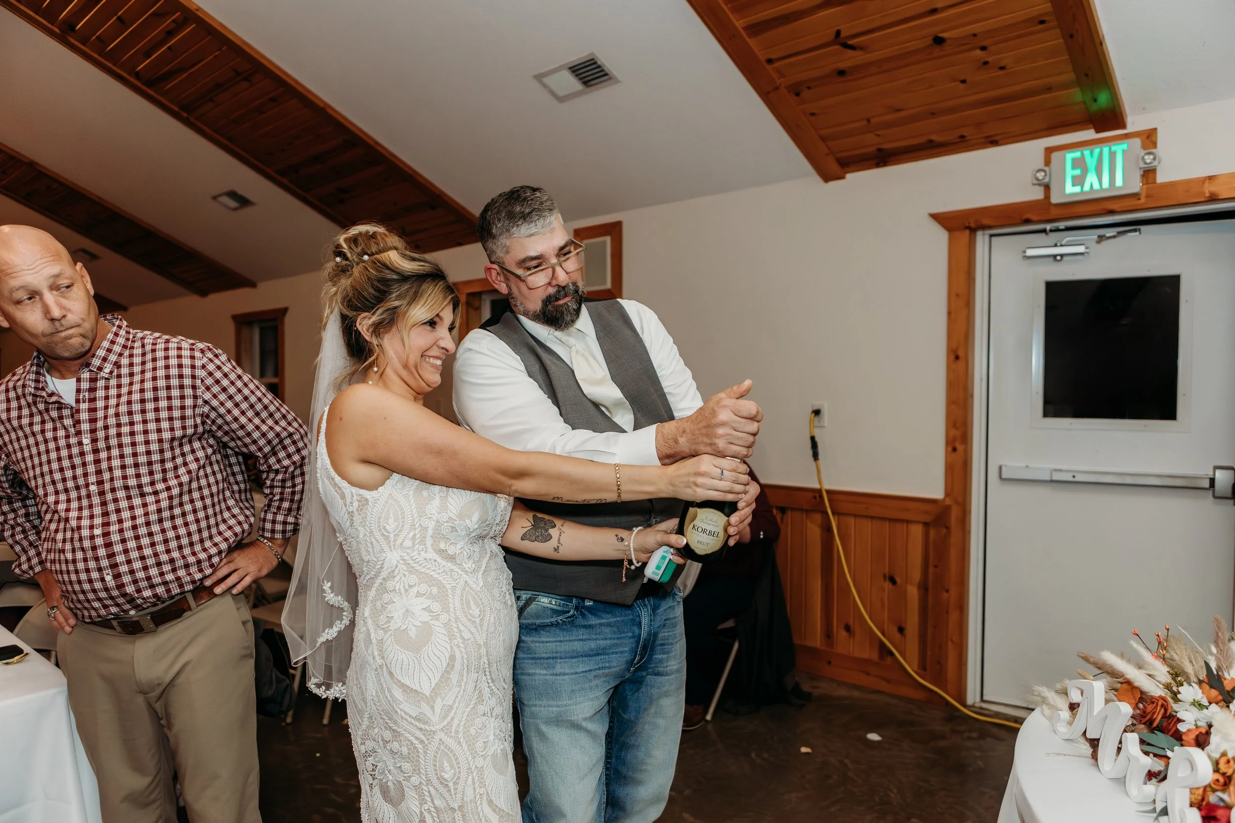 A bride and groom opening a bottle of champagne at a wedding reception, with a guest standing nearby and an exit sign above the door.