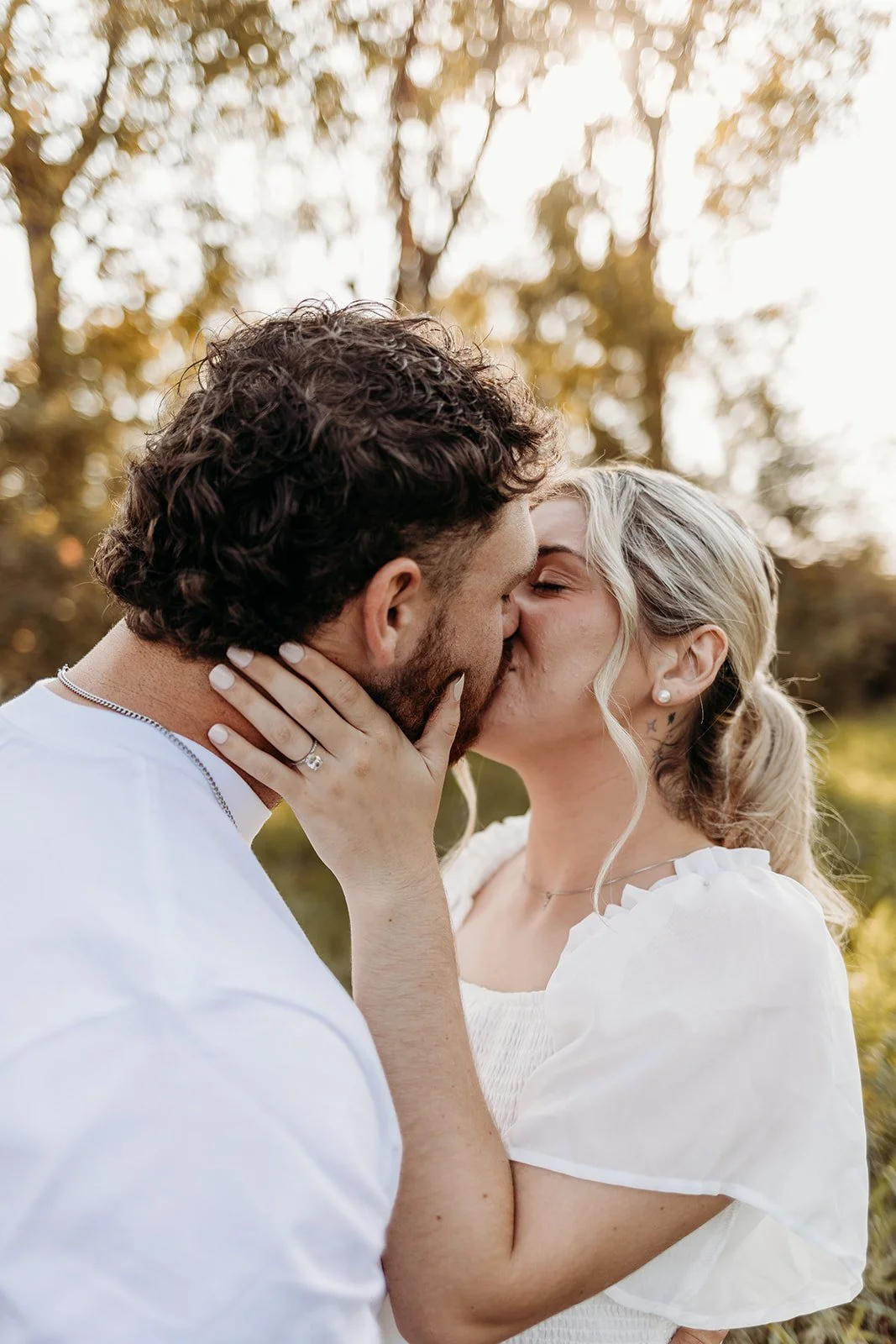 A couple sharing a kiss outdoors during sunset, with blurred trees in the background.