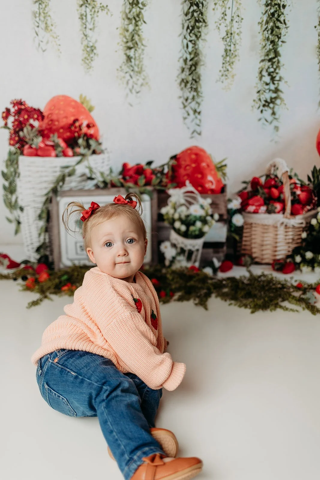 A young girl with blonde hair styled in two pigtails with red bows, wearing a pink sweater and jeans, sitting on the floor in front of a festive strawberry-themed decoration background with strawberries, flowers, and greenery.
