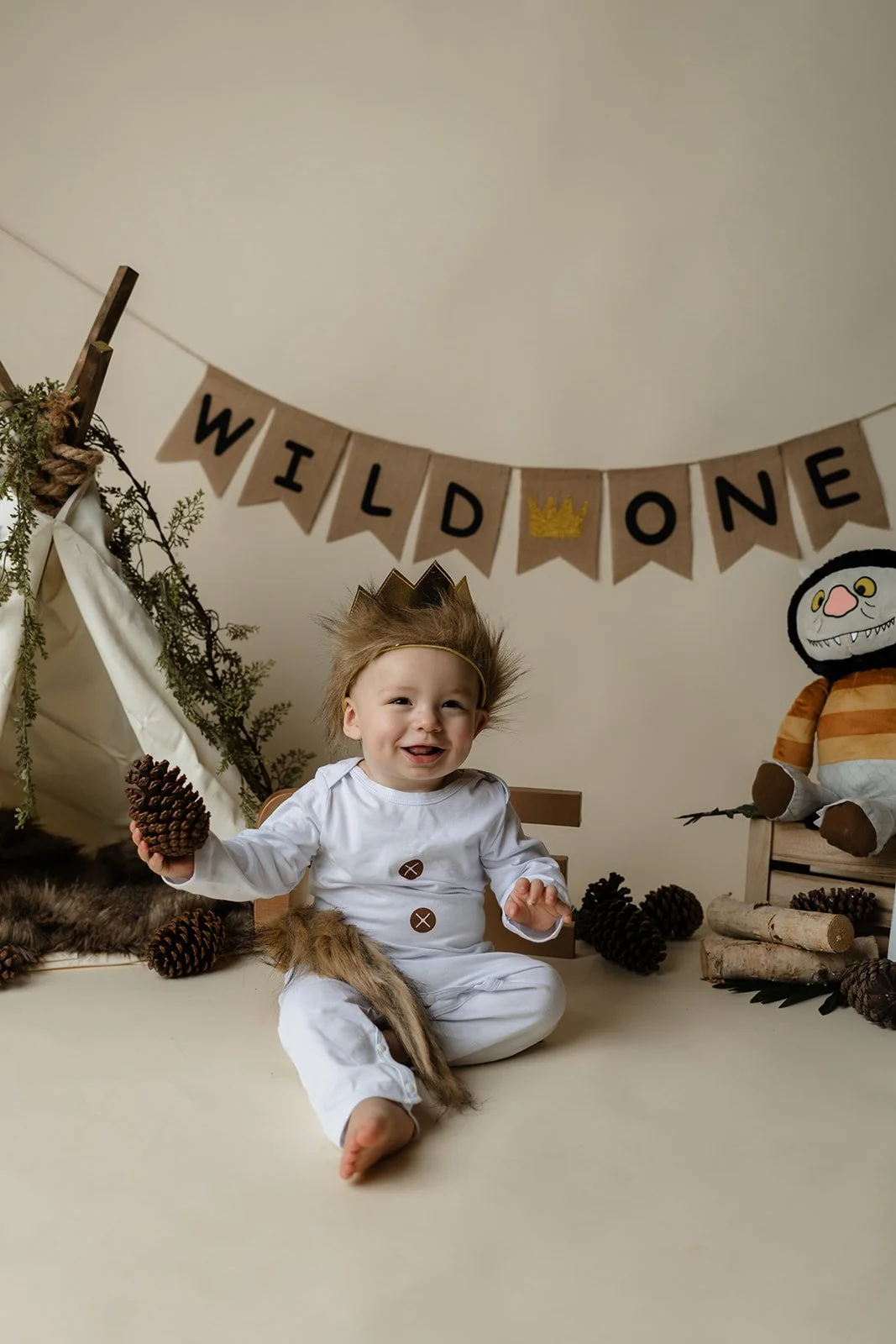A smiling child dressed as a caveman, sitting on the floor with pinecones around, a makeshift teepee to the left, and a plush monster to the right, in front of a 'WILD ONE' banner with a golden crown.