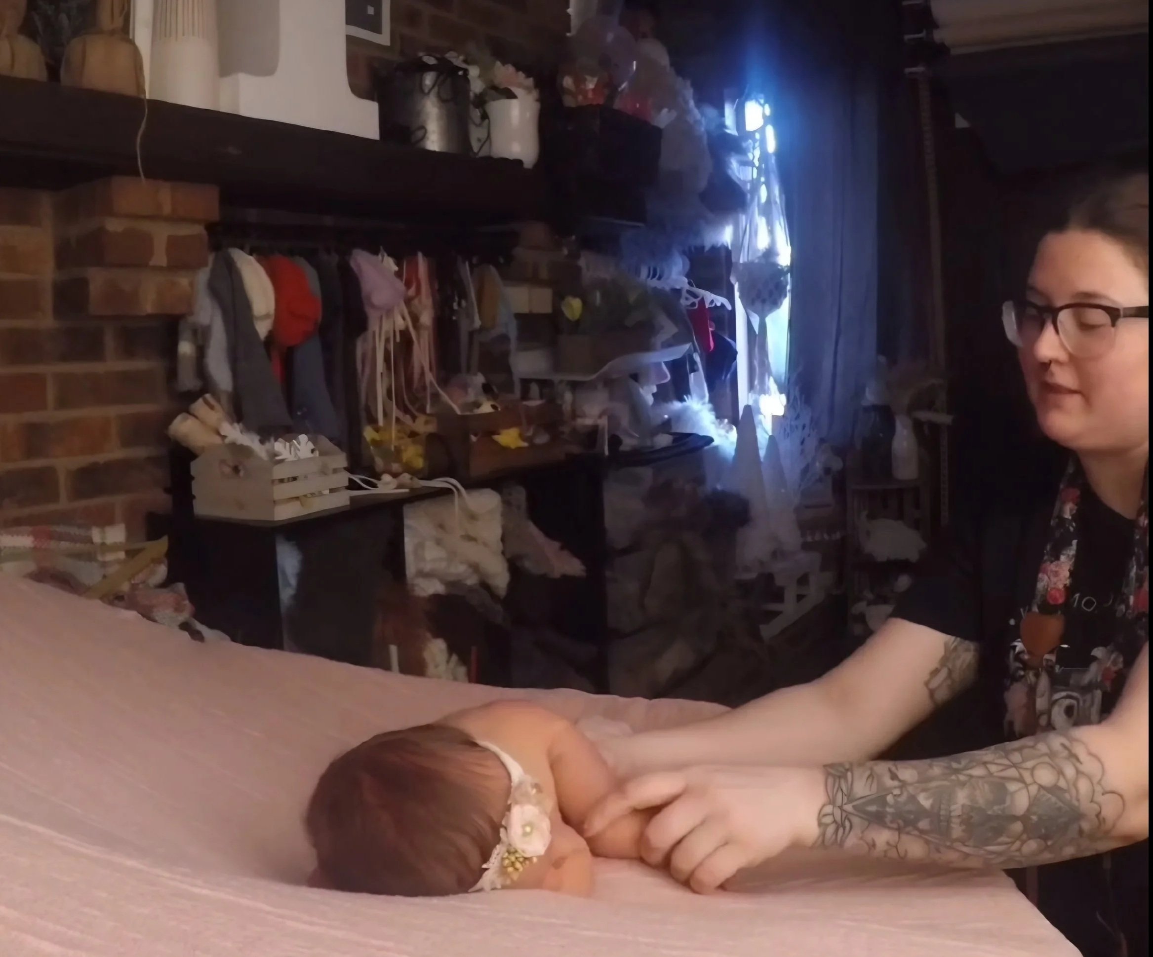 A woman with glasses and arms covered in tattoos gently touches the hand of a sleeping baby lying on a pink surface, wearing a floral headband, in a dimly lit room with a brick wall and shelves filled with various objects in the background.