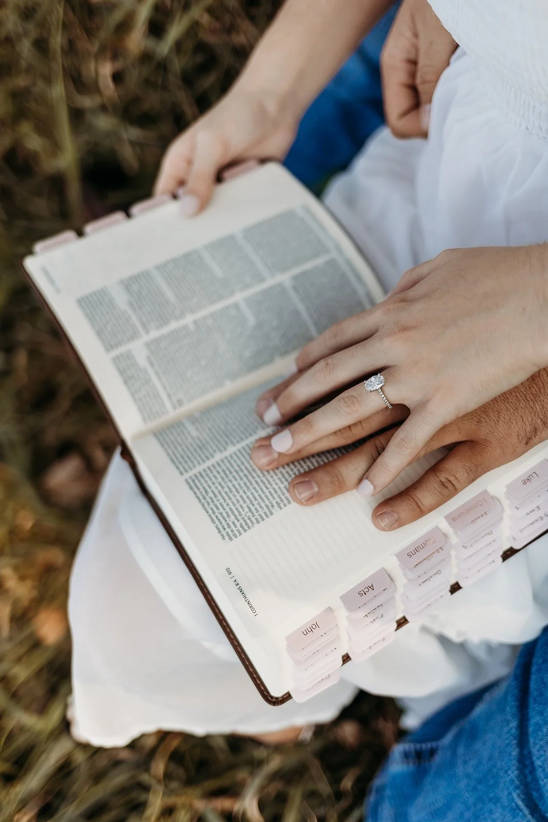 A person wearing a white dress and an engagement ring resting on a Bible open to a page with small text, outside on a grassy surface.