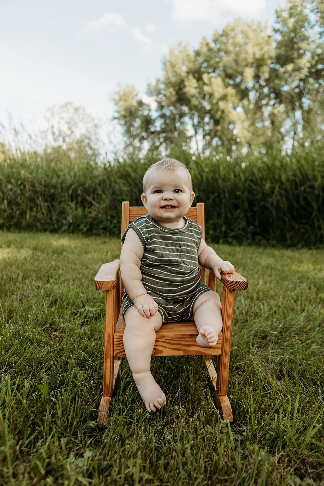 A baby sitting in a wooden chair outdoors on grass with trees and a blue sky in the background.