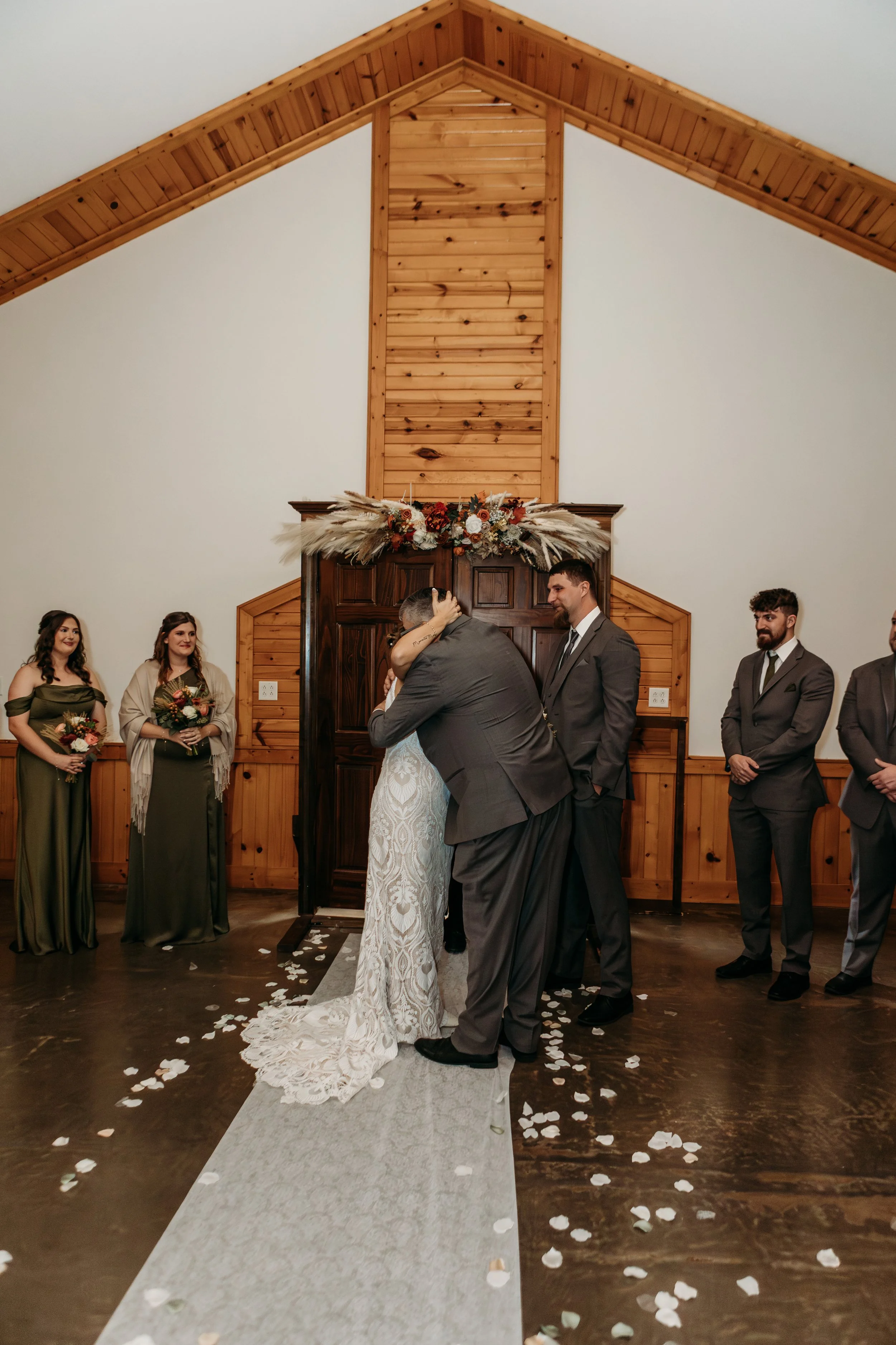 A bride and groom share a kiss during their wedding ceremony in a rustic indoor setting with wooden walls and a sloped ceiling. Bridesmaids and groomsmen stand on either side, with flower petals scattered on the floor.