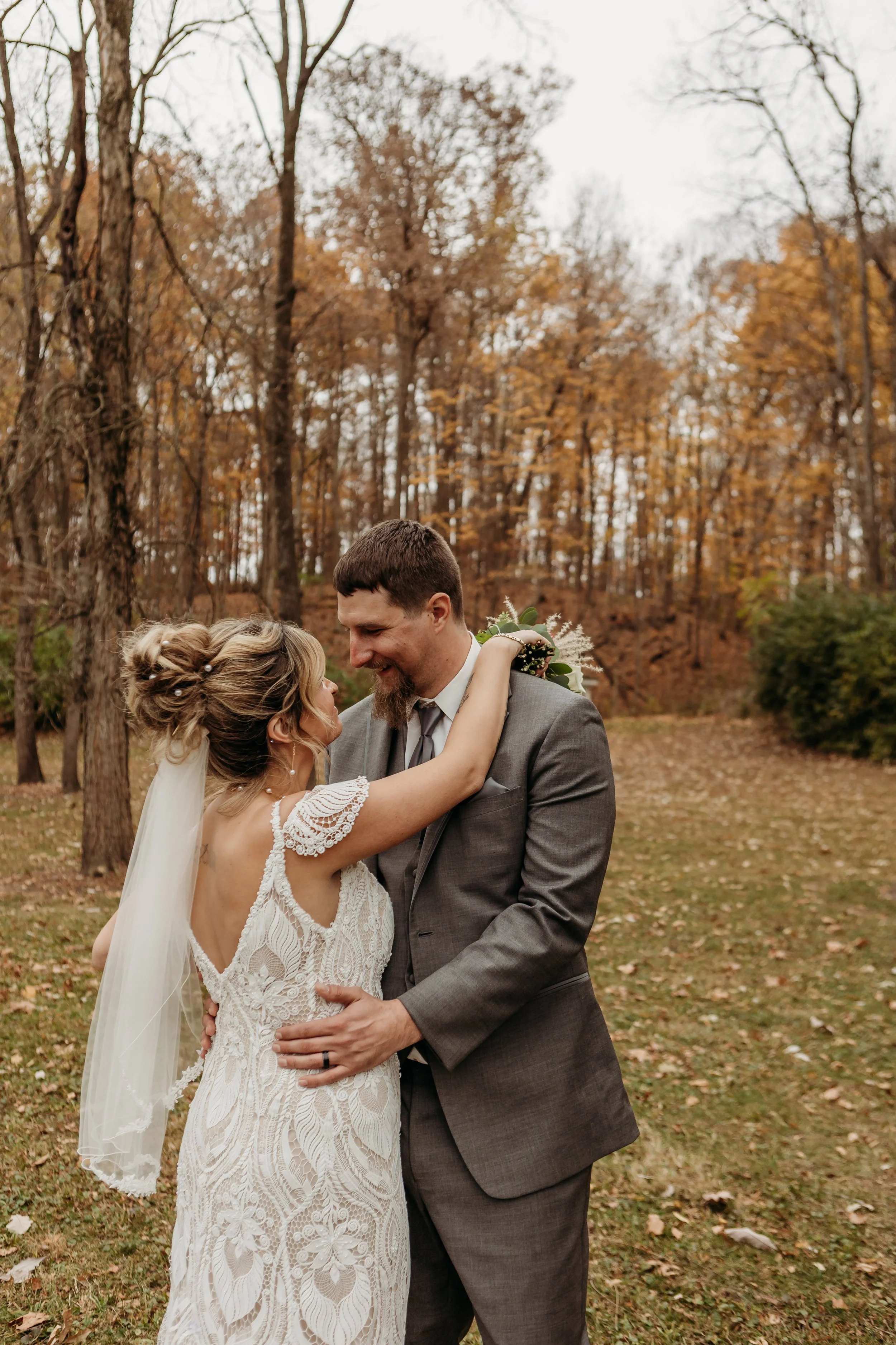 A bride and groom embrace outdoors in autumn, surrounded by fall-colored trees, during their wedding.