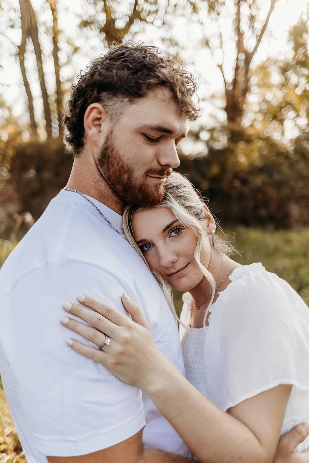 A couple cuddling outdoors during autumn, with the woman resting her head on the man's chest, showing her engagement ring.
