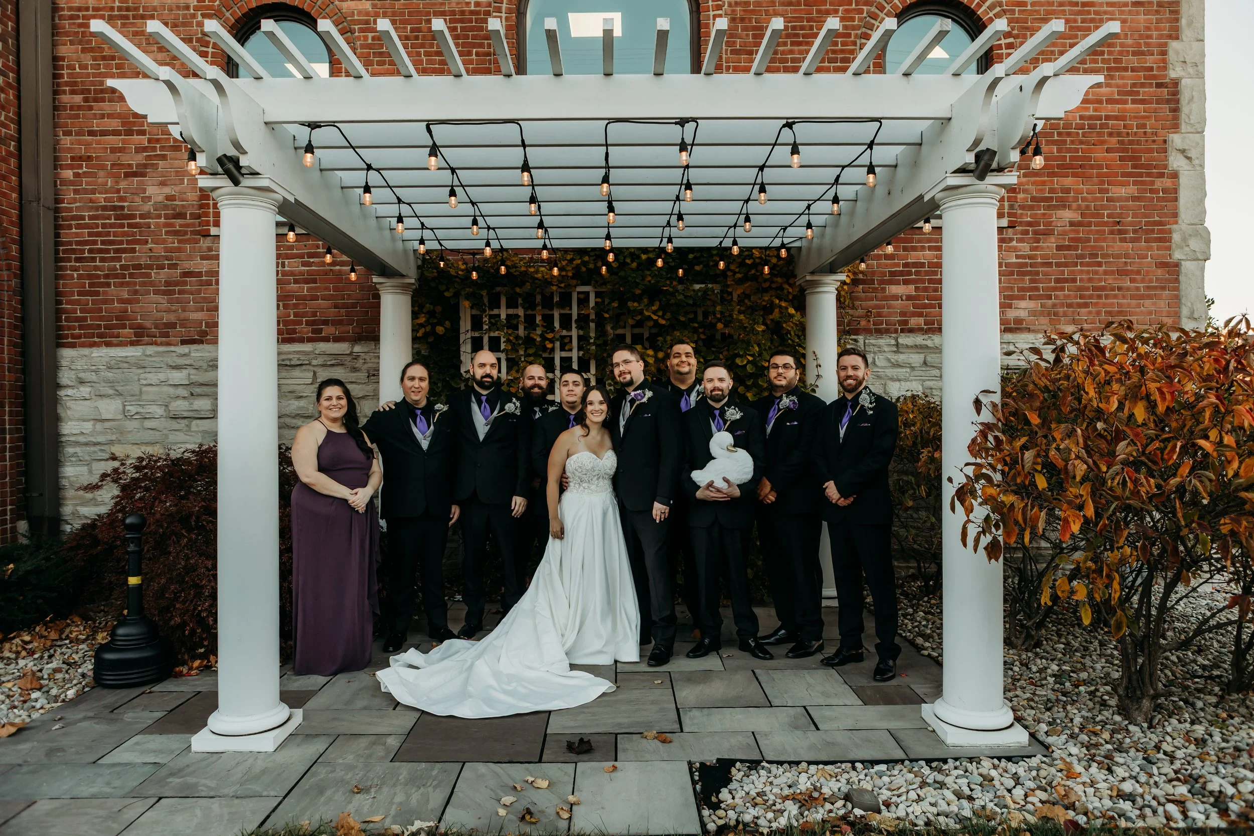 Wedding party photo under a pergola with string lights, featuring a bride in a white wedding dress, groom, and several men in suits, with a woman in a purple dress on the side, set against a brick and stone building with autumn foliage.