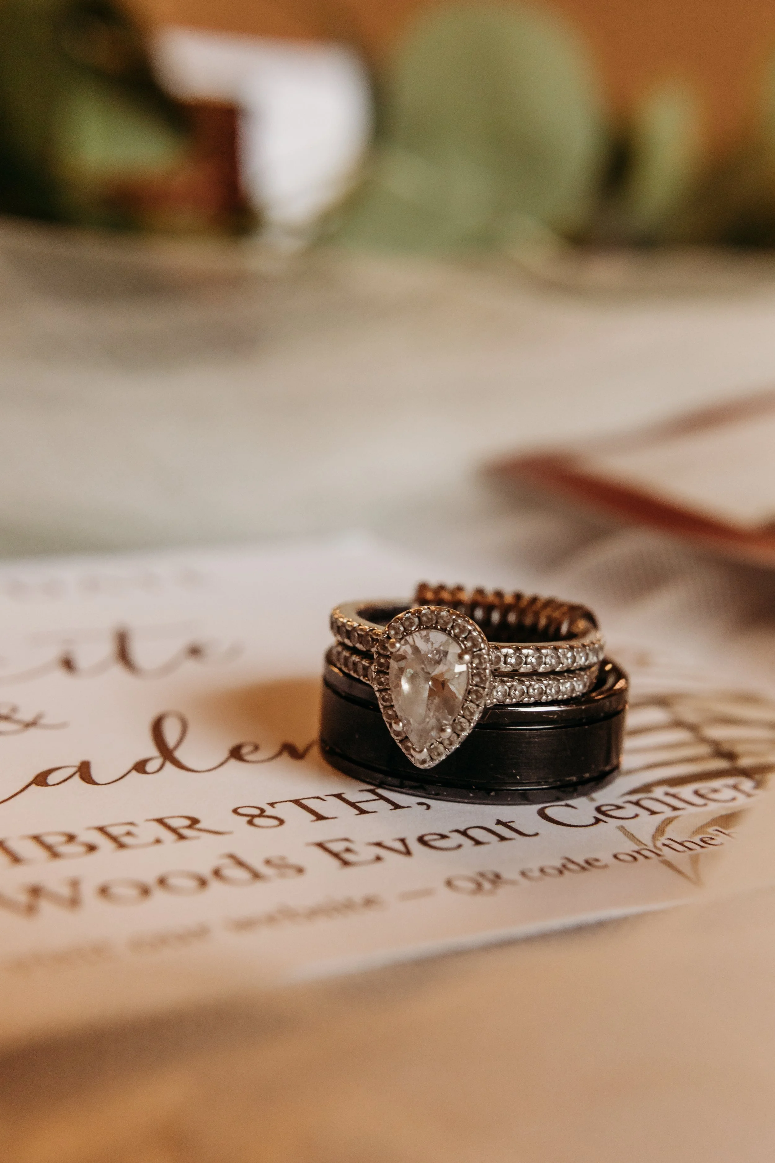 A close-up of a pear-shaped diamond ring surrounded by smaller diamonds, set on top of stacked rings, resting on an event card.
