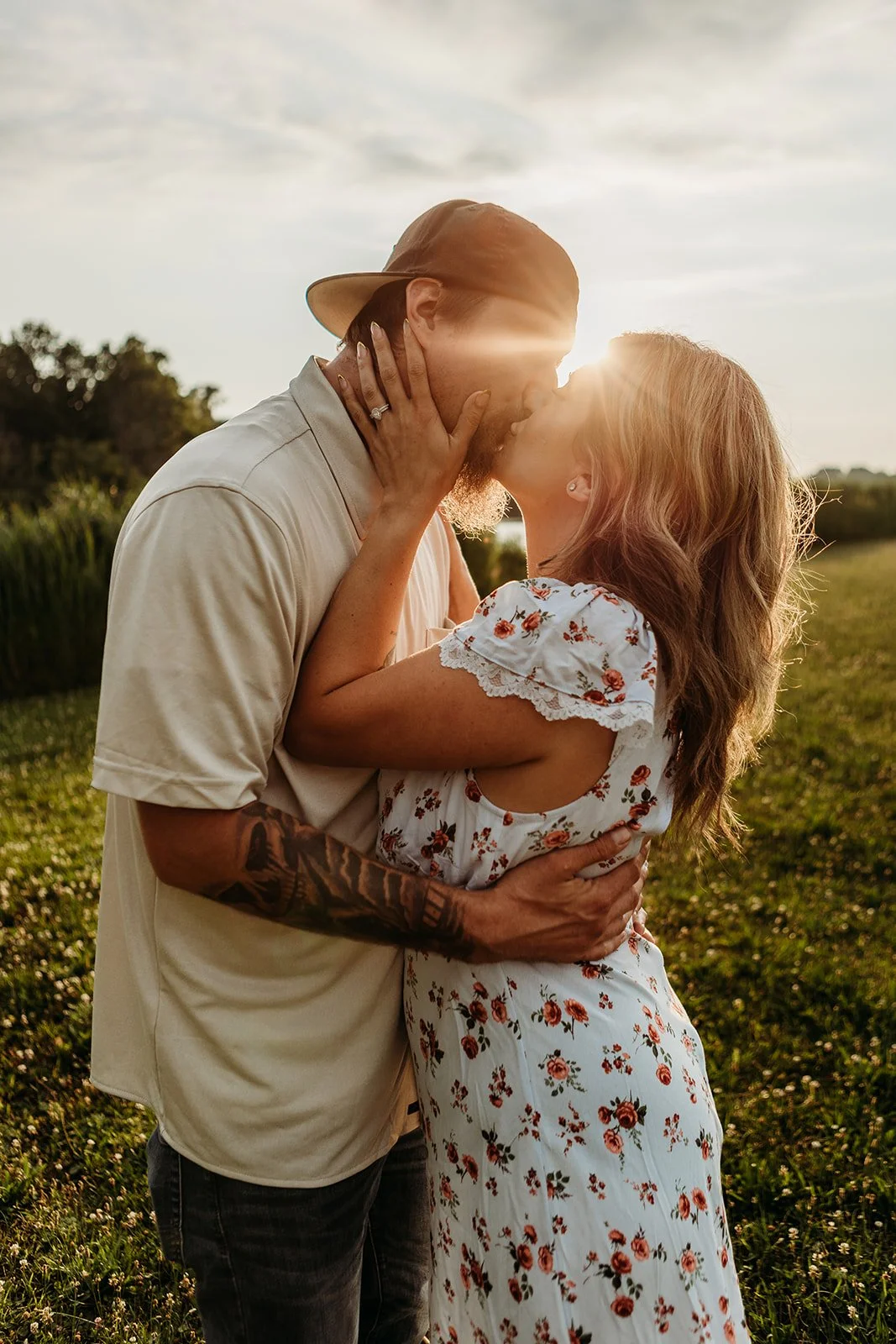 A couple kissing outdoors during sunset, with the man wearing a beige shirt and a cap, and the woman in a white floral dress. The woman has her hand on the man's face, and the man has his arm around her waist, holding her close.