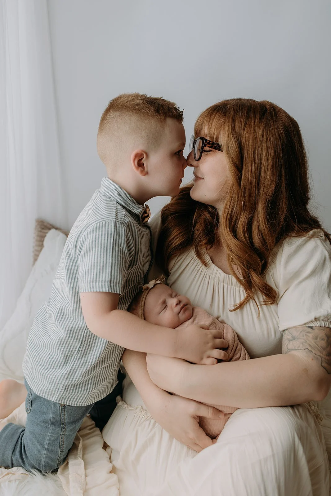 A woman with red hair and glasses holding a sleeping baby girl, while a young boy leans in to touch noses with her, in a cozy, softly lit room.