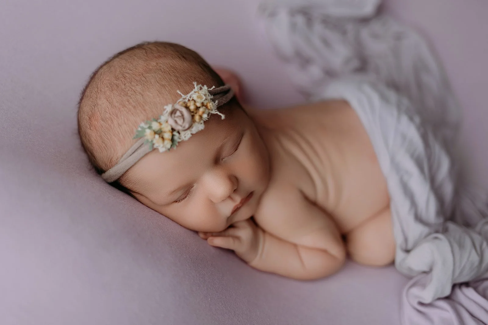 A sleeping newborn baby wearing a floral headband, lying on a soft pink surface with a relaxed posture, with arms bent and hands near the face.