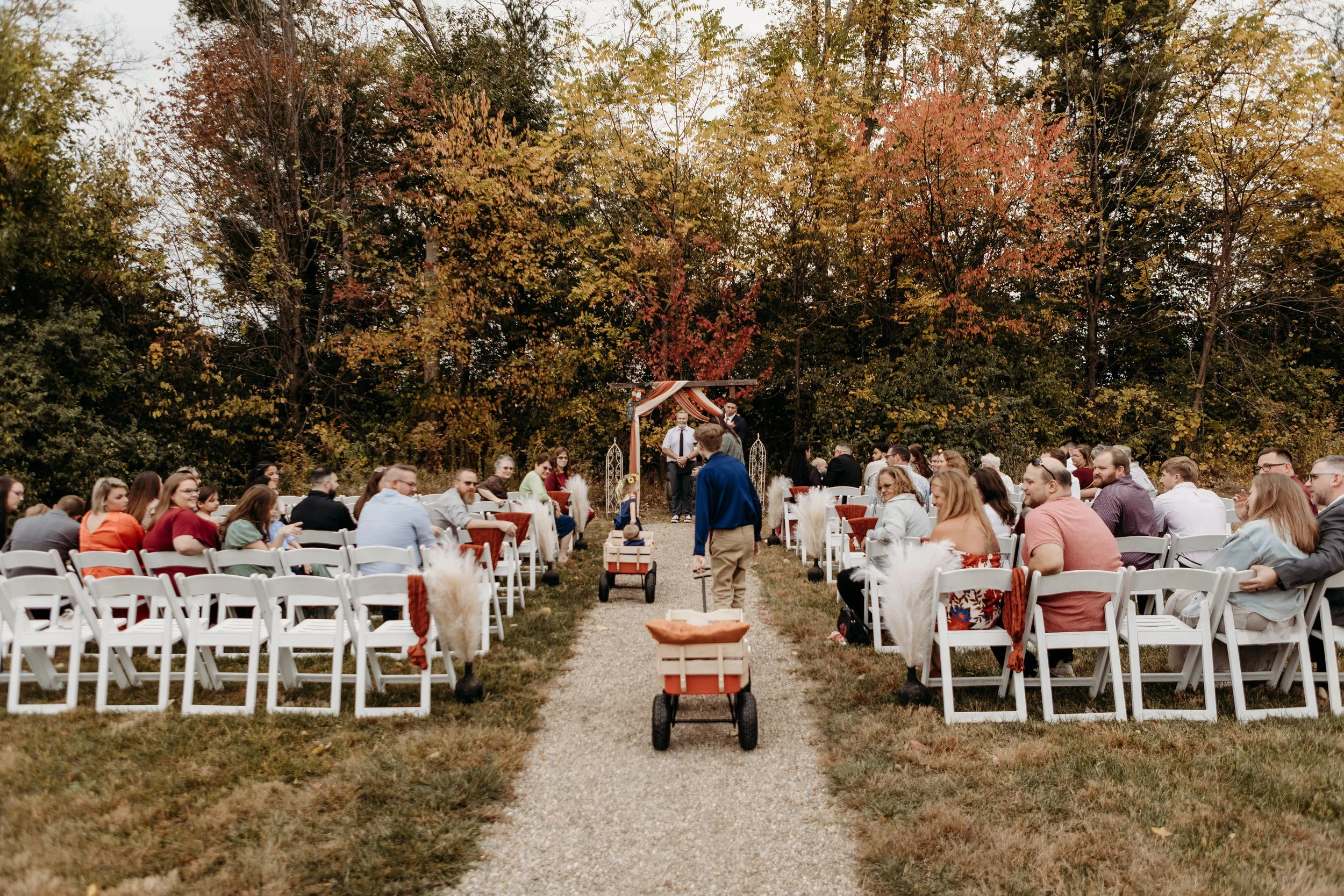 Outdoor wedding ceremony with guests seated on white chairs on either side of a gravel aisle, with a couple standing at the altar under a decorated canopy, surrounded by fall trees with colorful autumn leaves.
