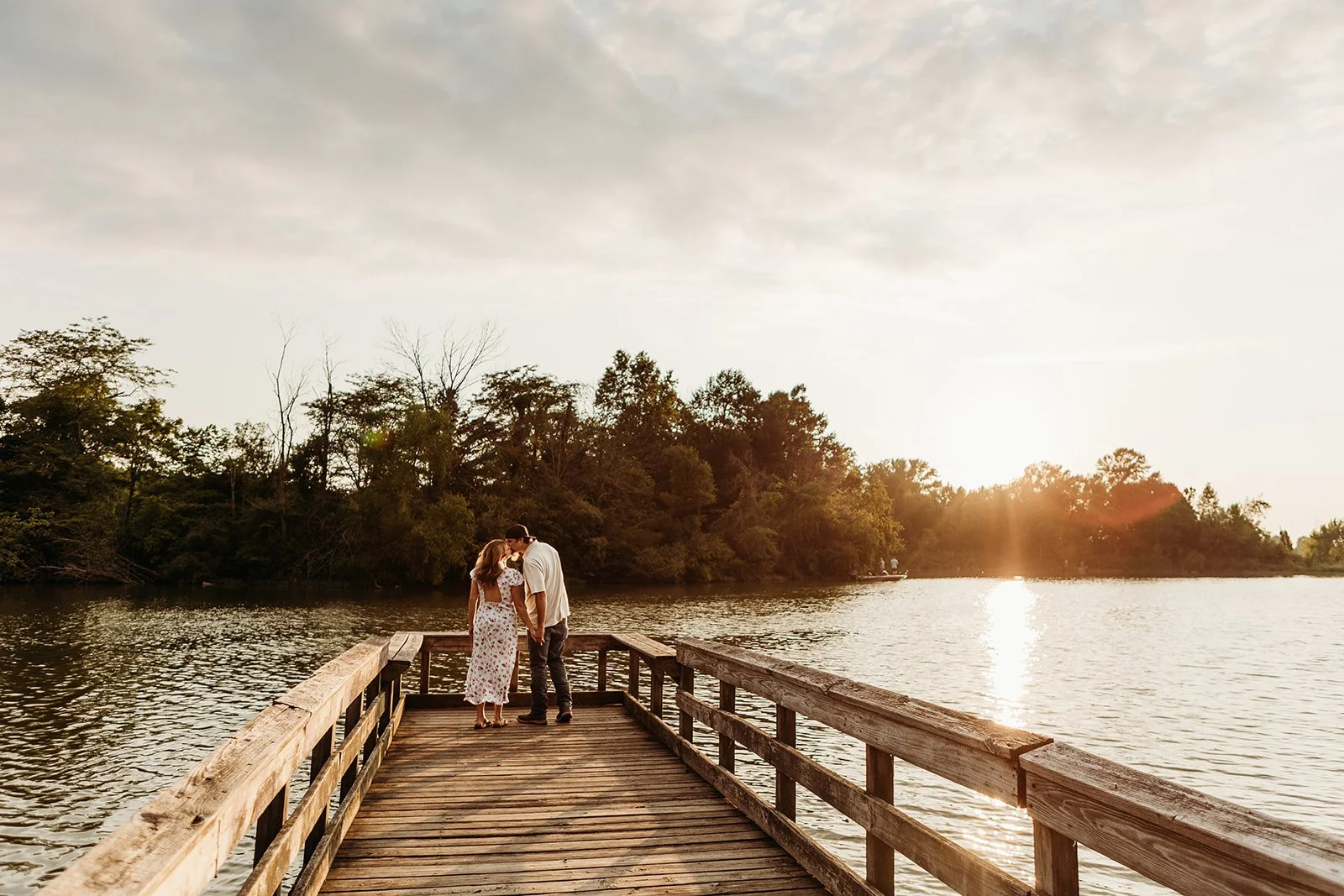 A couple stands on a wooden dock by a lake at sunset, close together and kissing, with trees in the background and the sun low in the sky.