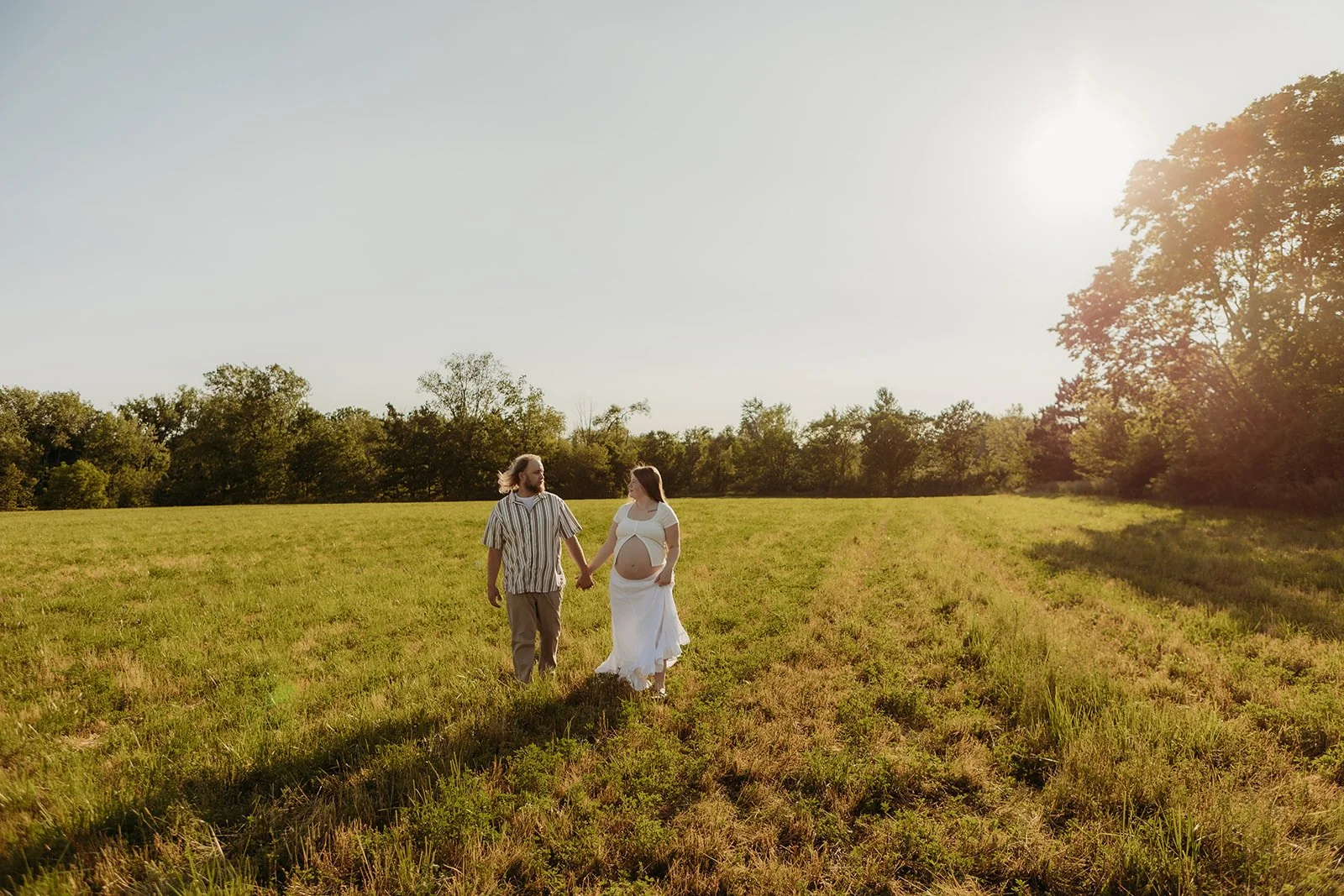 A pregnant woman and her partner holding hands while walking through a grassy field during sunset.