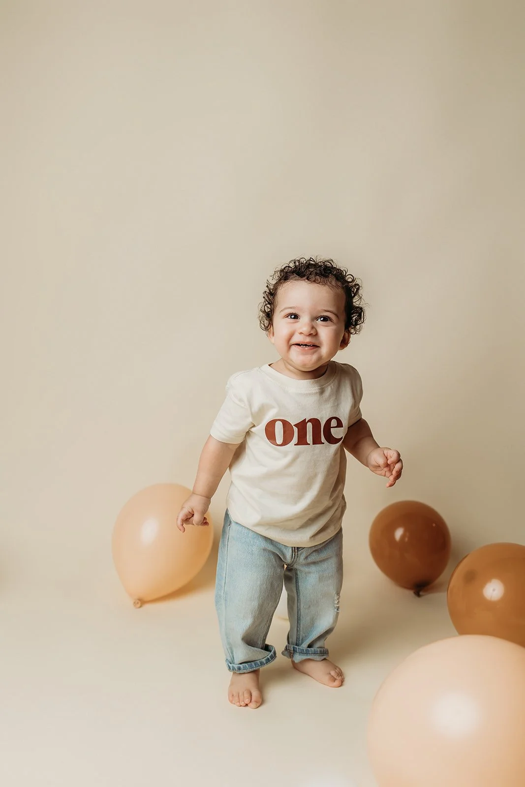 Adorable toddler with curly hair smiling, wearing a cream 'one' t-shirt and light denim pants, standing barefoot among beige and brown balloons against a simple beige background.