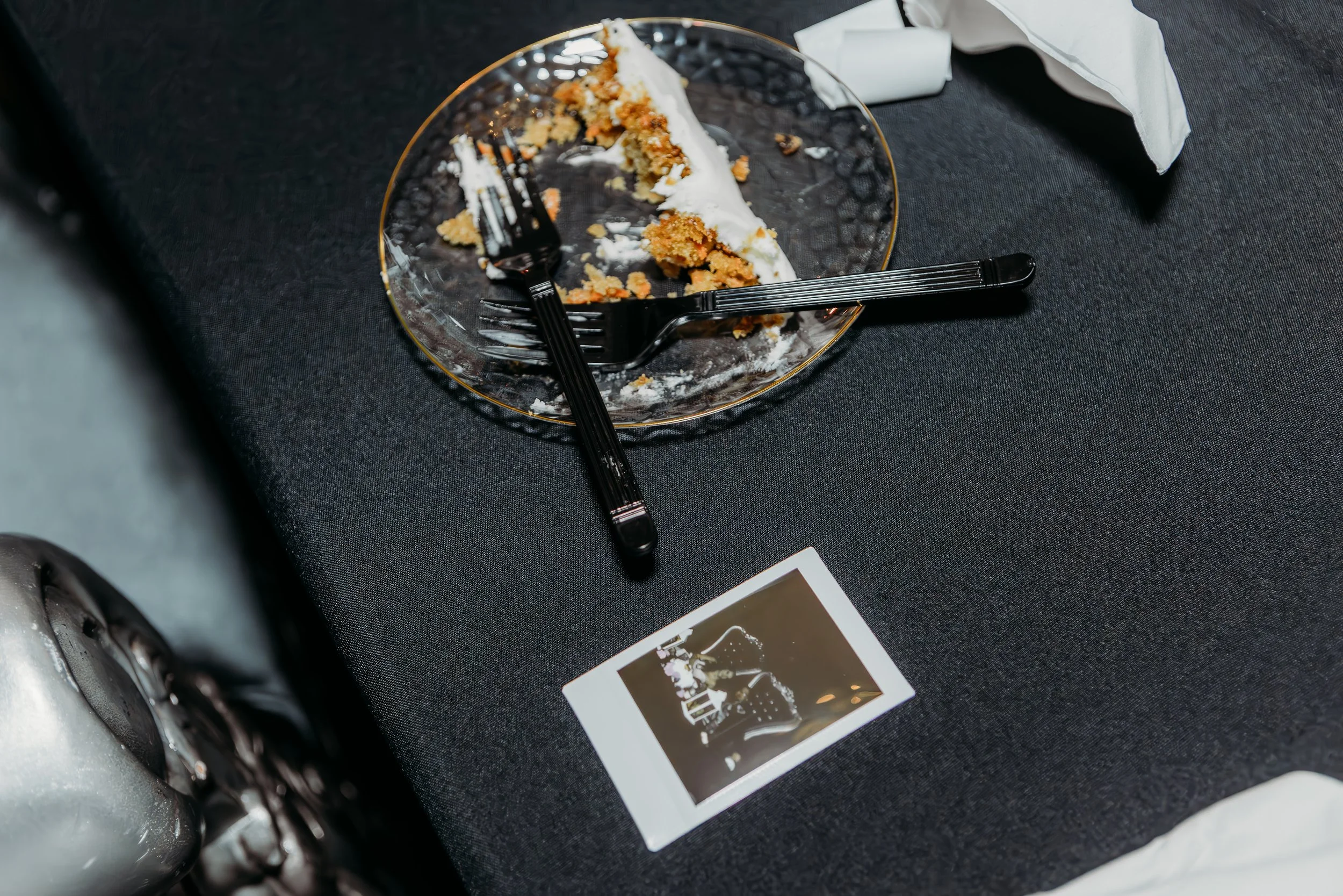 A black platter with a partially eaten slice of cake, two black forks placed on the plate, a crumpled white napkin, and a small printed photo on a black tablecloth.