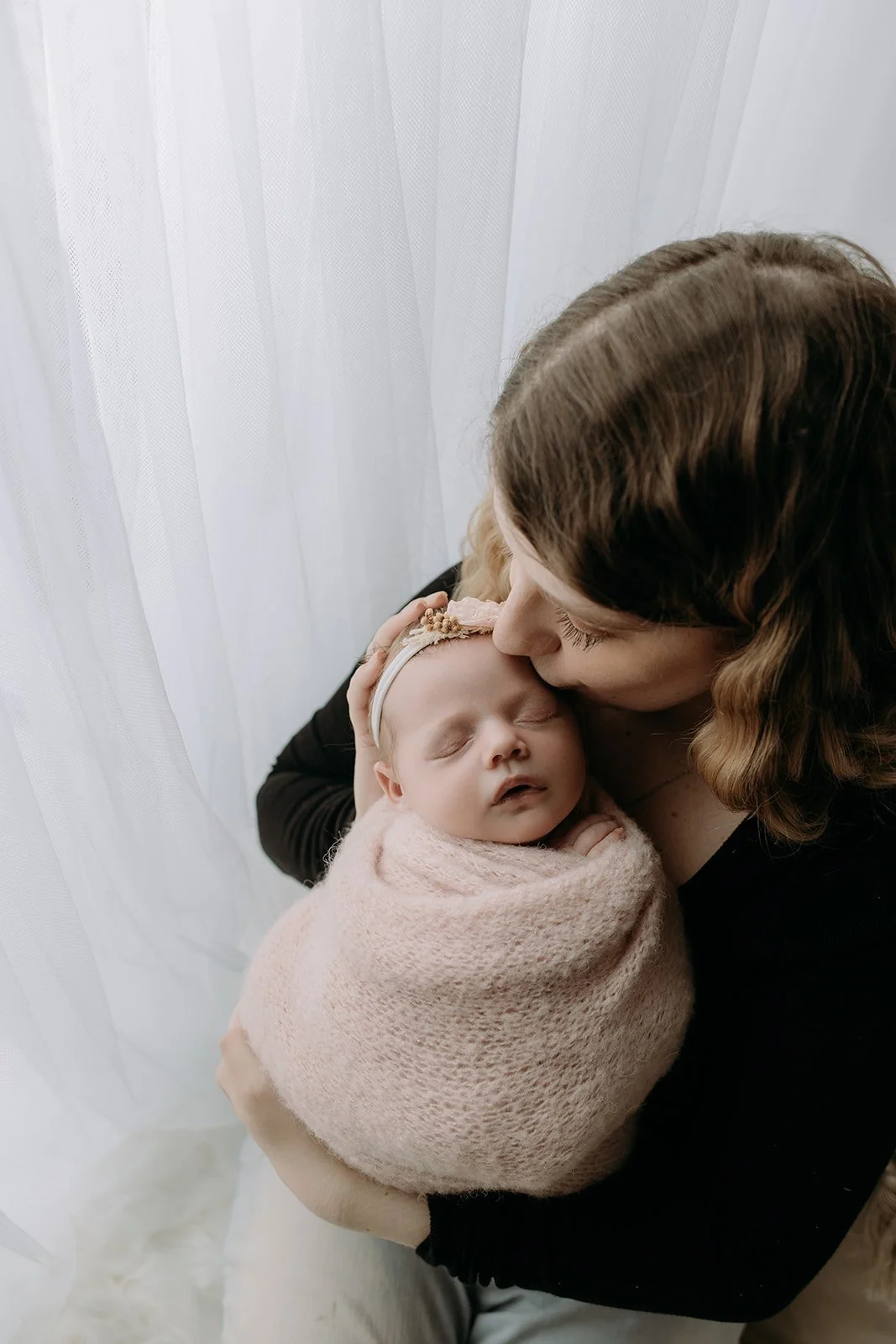 A woman holding a sleeping baby wrapped in a pink blanket near a white curtain.