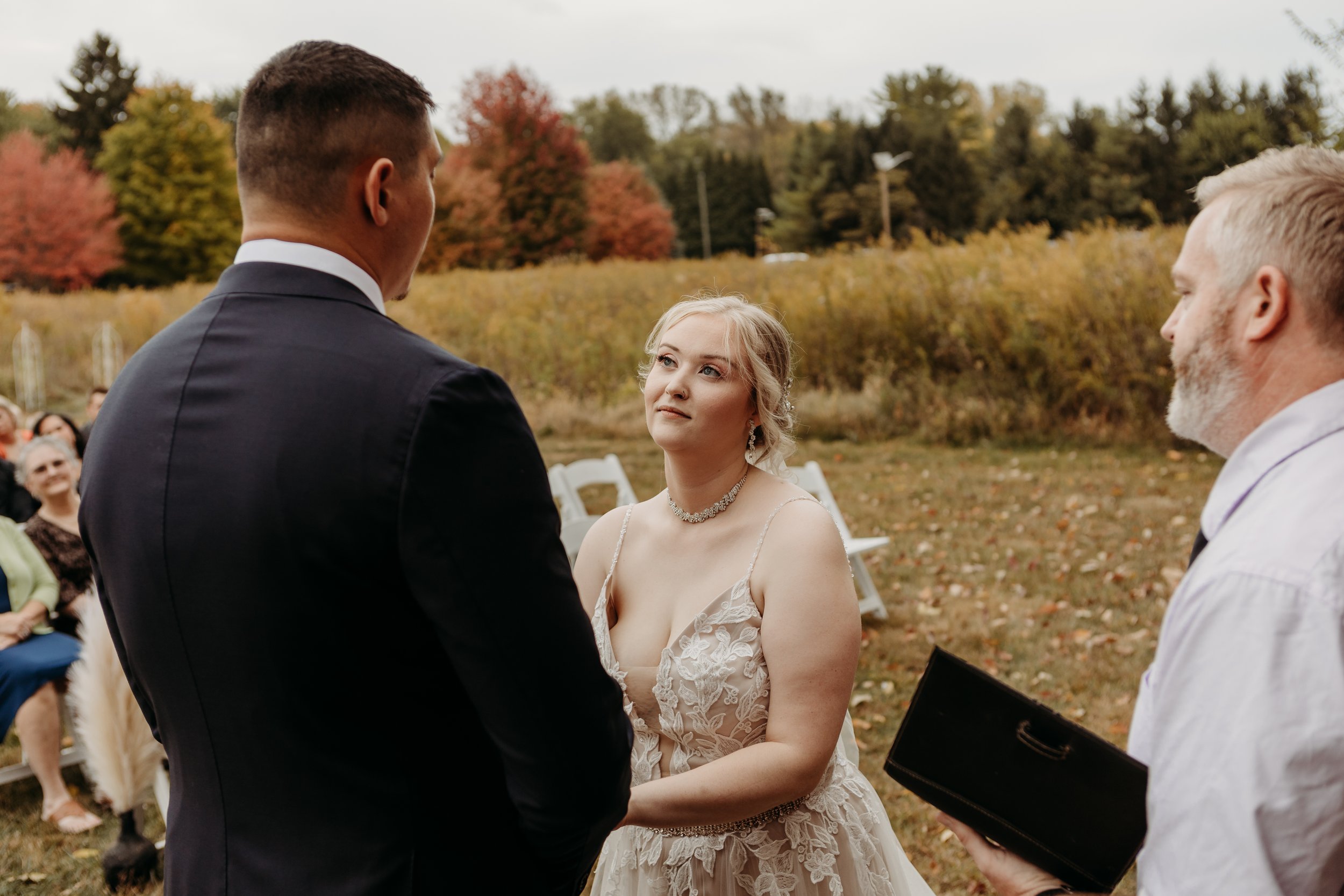 A bride and groom exchange vows outdoors during a fall wedding ceremony, with guests seated in the background and colorful autumn trees.