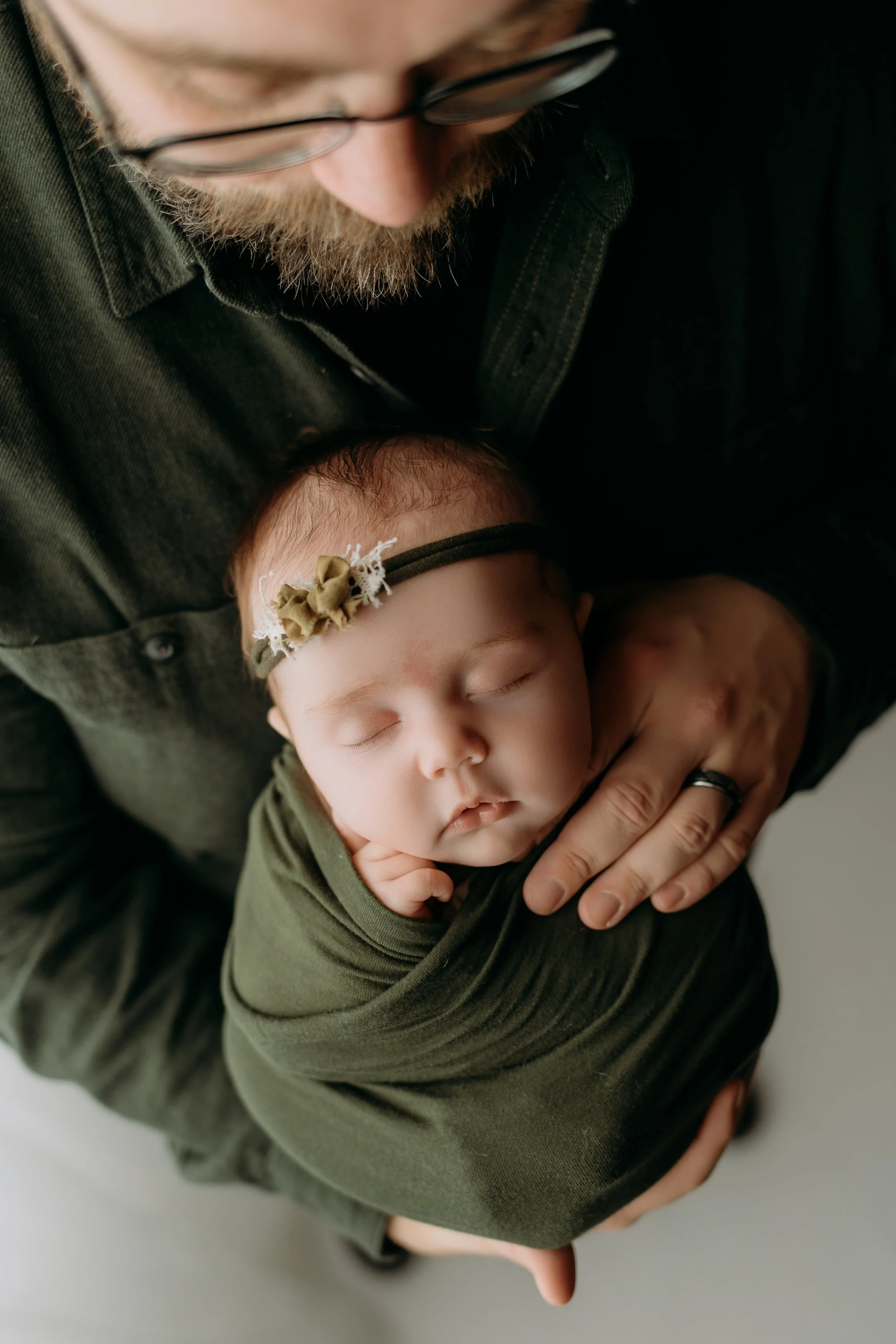 A man holding a sleeping baby wrapped in a green blanket, with the baby wearing a matching green headband with a small fabric flower.