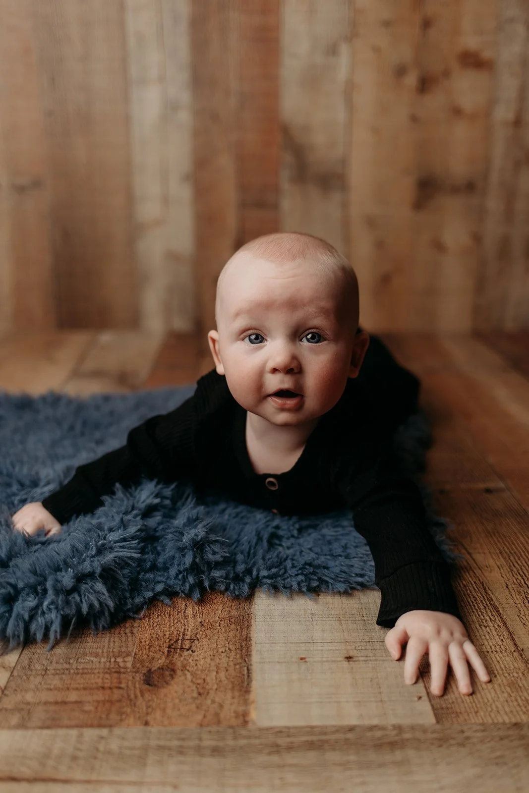 A baby with blue eyes crawling on a wooden floor, wearing a black outfit, on a blue furry blanket with a wooden background.