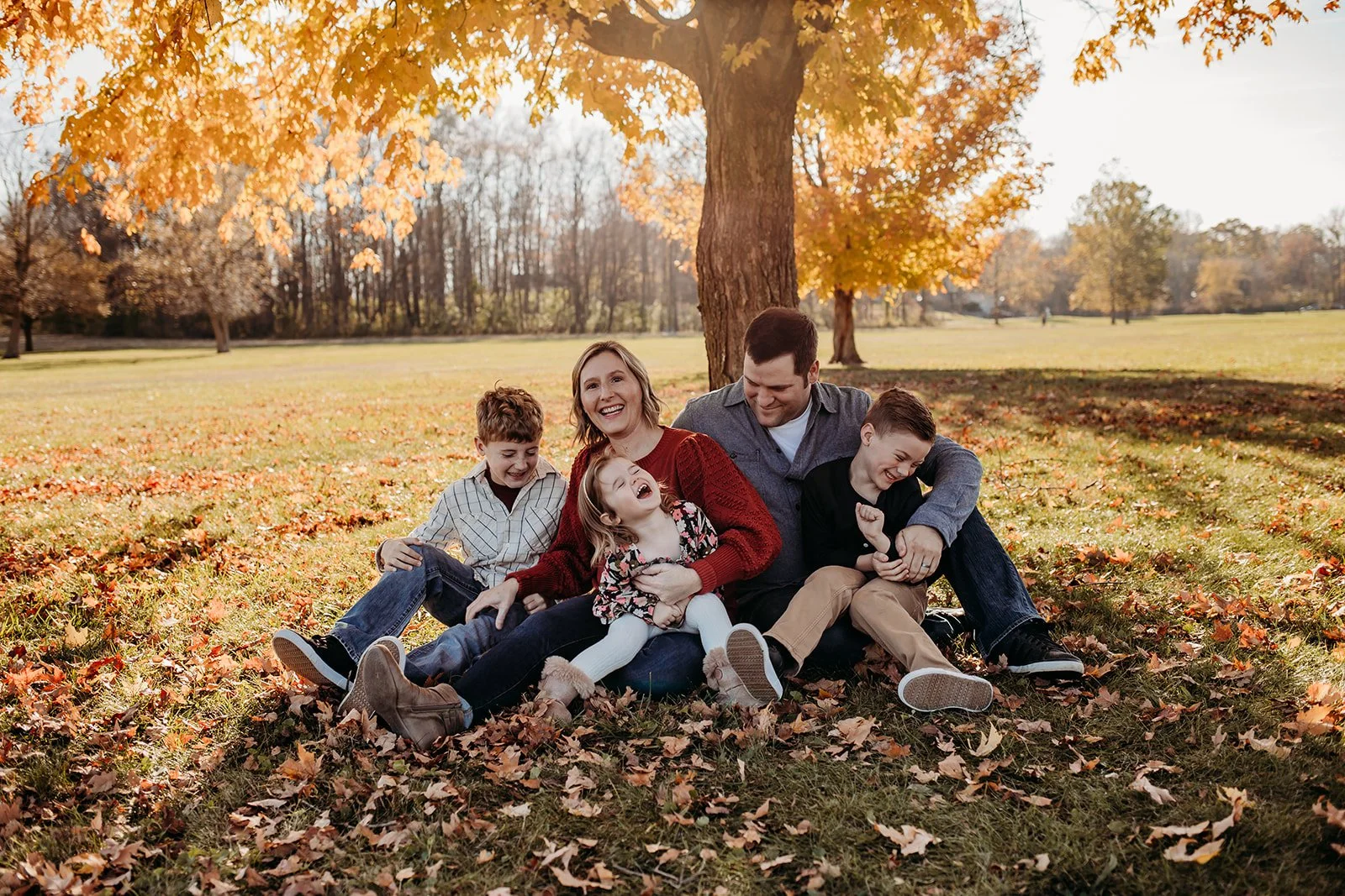 A family of six sitting on the ground in a park during fall, laughing and playing together near a tree with orange leaves.