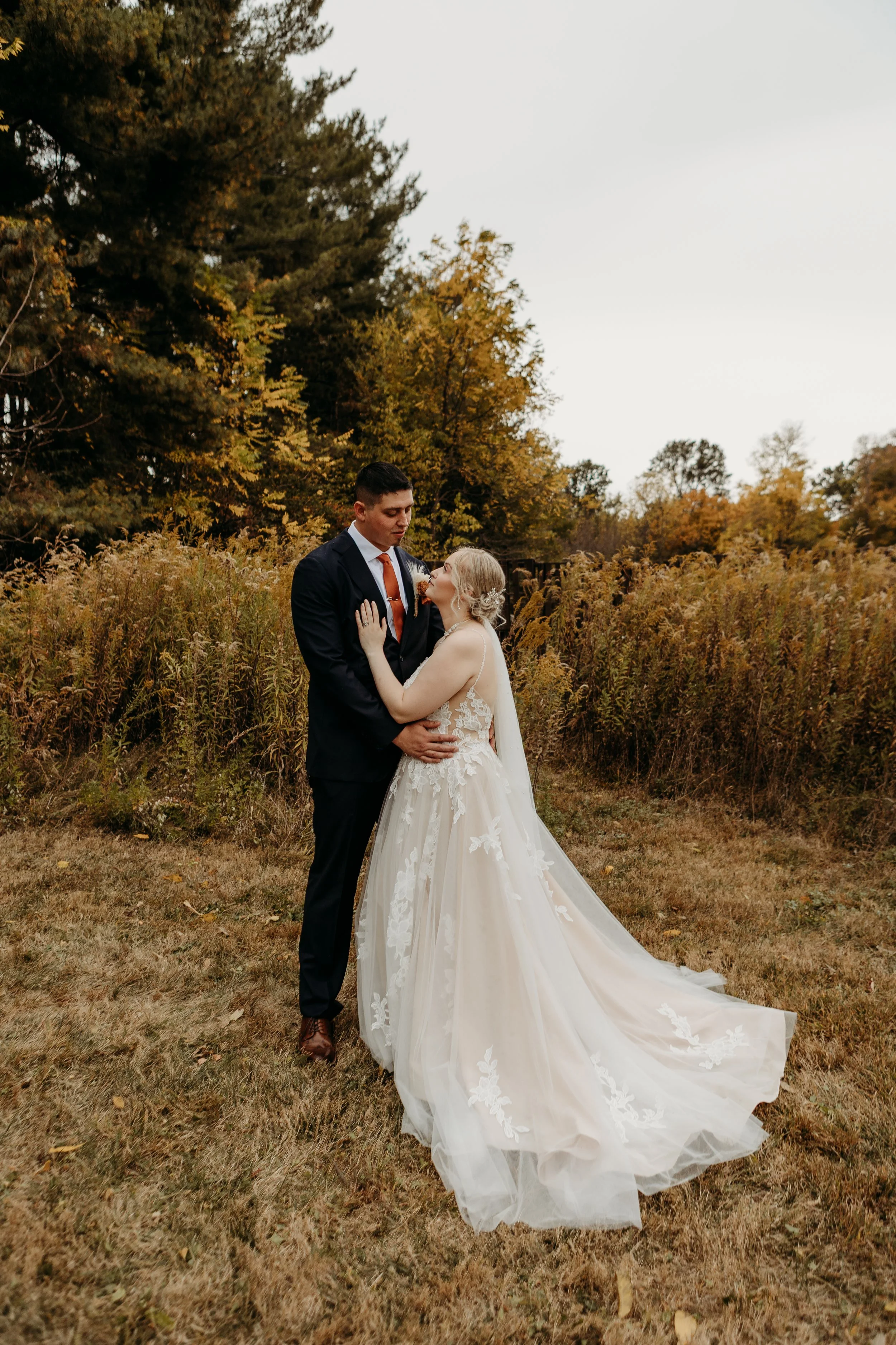 A bride and groom standing close together outdoors, surrounded by trees with fall foliage, on a grassy area during daytime.