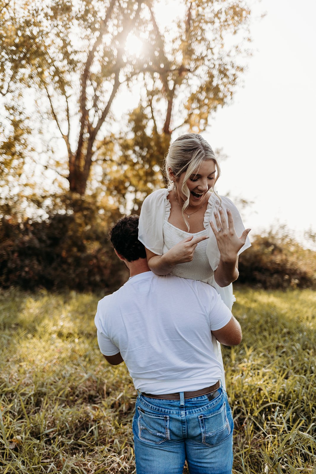 A joyful woman celebrates her engagement ring while being carried on a man's shoulders outdoors during sunset, with trees and grass in the background.