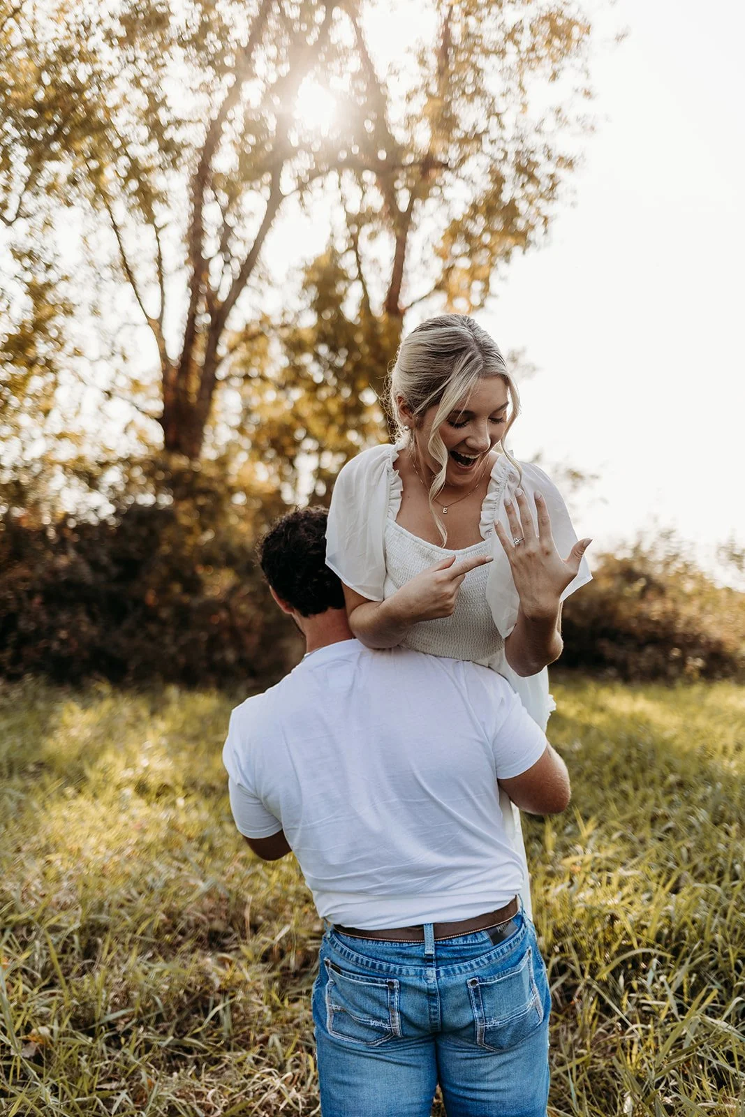 A joyful woman celebrates her engagement ring while being carried on a man's shoulders outdoors during sunset, with trees and grass in the background.