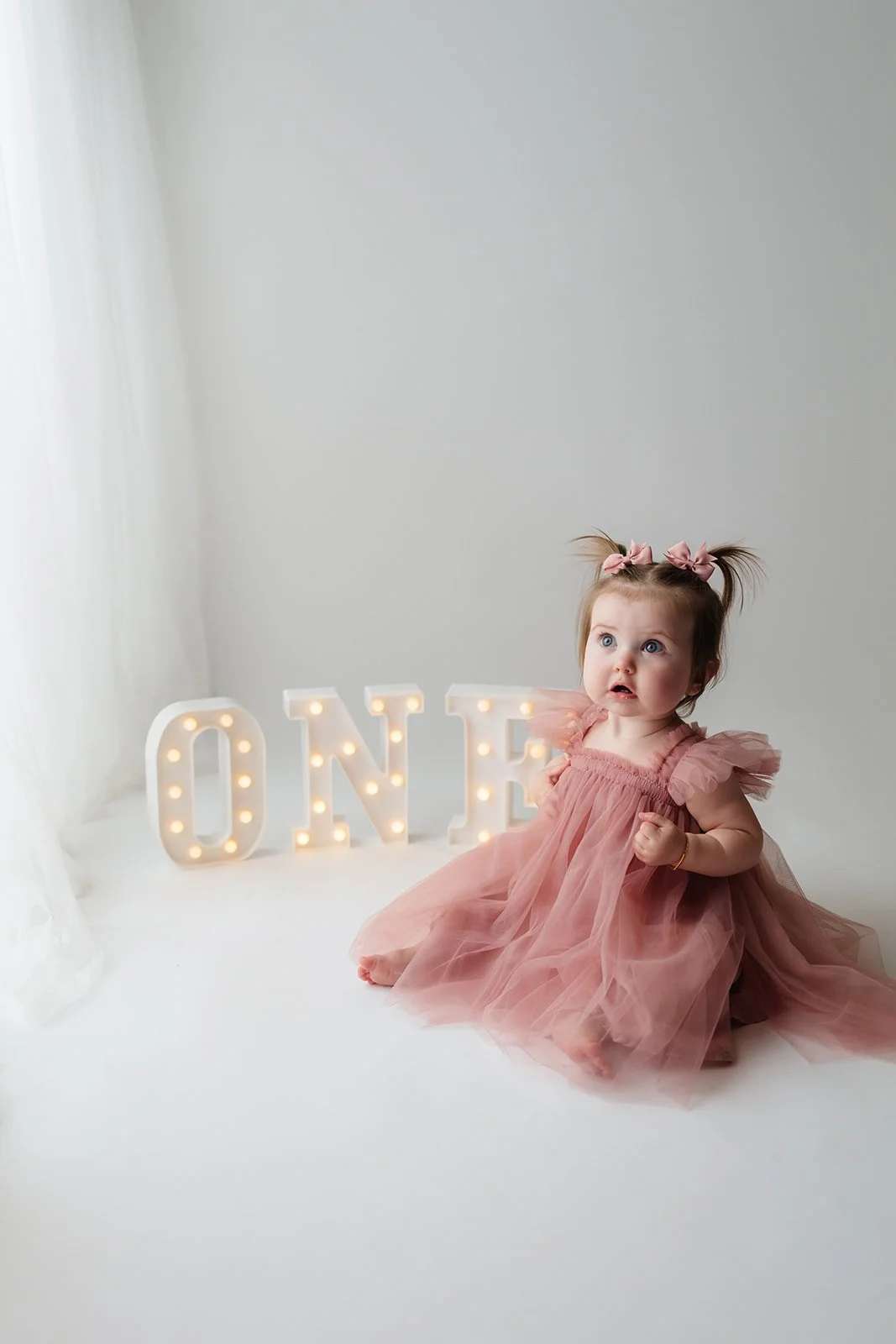 A young girl with blue eyes and light brown hair in pigtails, wearing a pink tulle dress with ruffled sleeves, sitting on a white surface. Behind her are illuminated marquee letters spelling 'ONE' on a light gray wall.