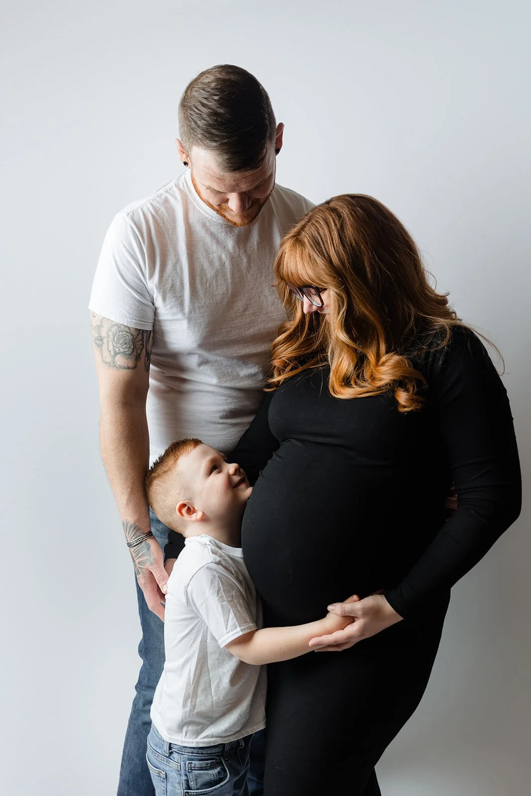 A pregnant woman with red hair and glasses gently holding her belly, a small boy hugging her from the front, and a man looking down at her, all smiling and sharing an affectionate moment.
