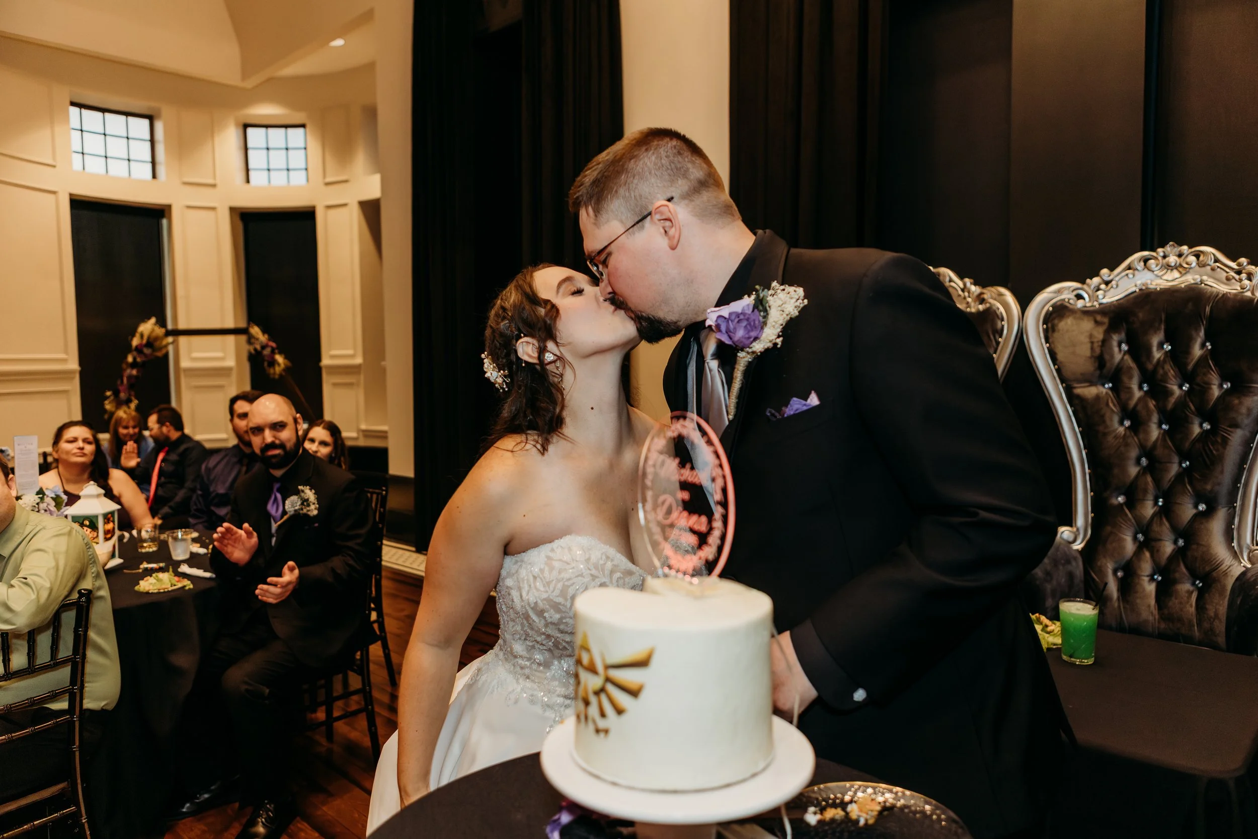A bride and groom kiss in front of a wedding cake at their wedding reception, with seated guests clapping in the background.