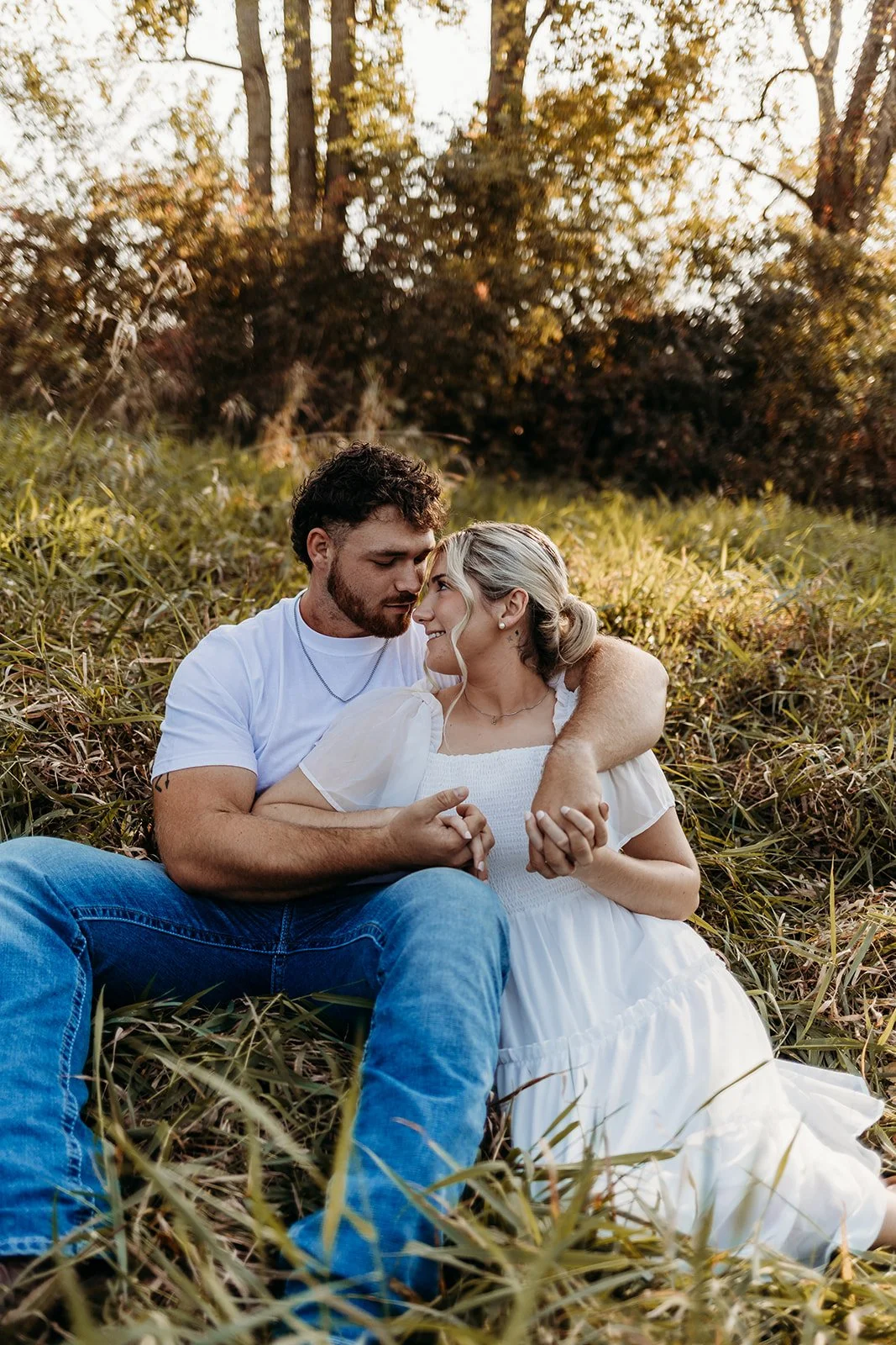 A couple sitting on grass in a natural outdoor setting during sunset, hugging and touching foreheads, looking into each other's eyes.