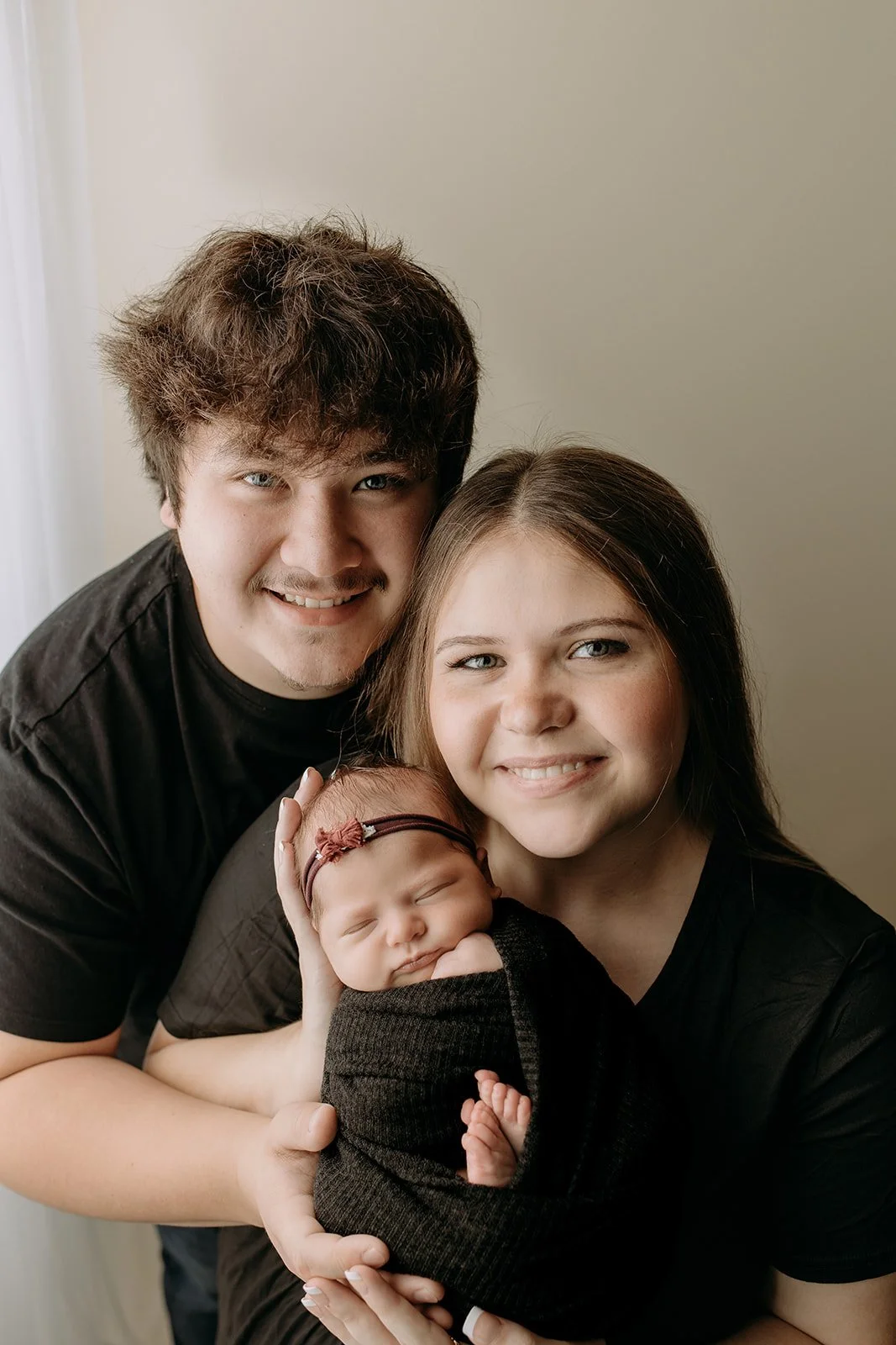A family of three is posing for a photo. The father is on the left with curly hair, the mother on the right with straight hair, and in the middle, they are holding a newborn baby wrapped in a black blanket. The baby has a rose headband and is sleepin