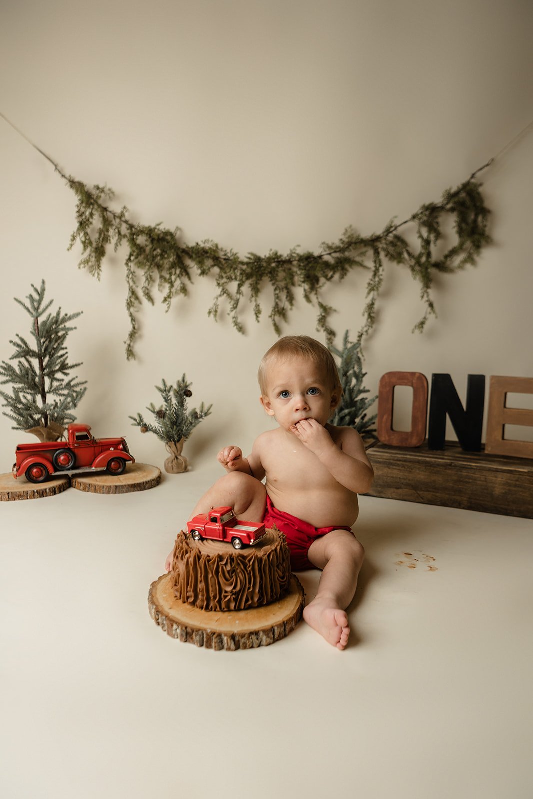 A baby sitting on a wooden slice, with a small toy truck on it, surrounded by Christmas decorations including mini pine trees and a garland, in a festive setting.