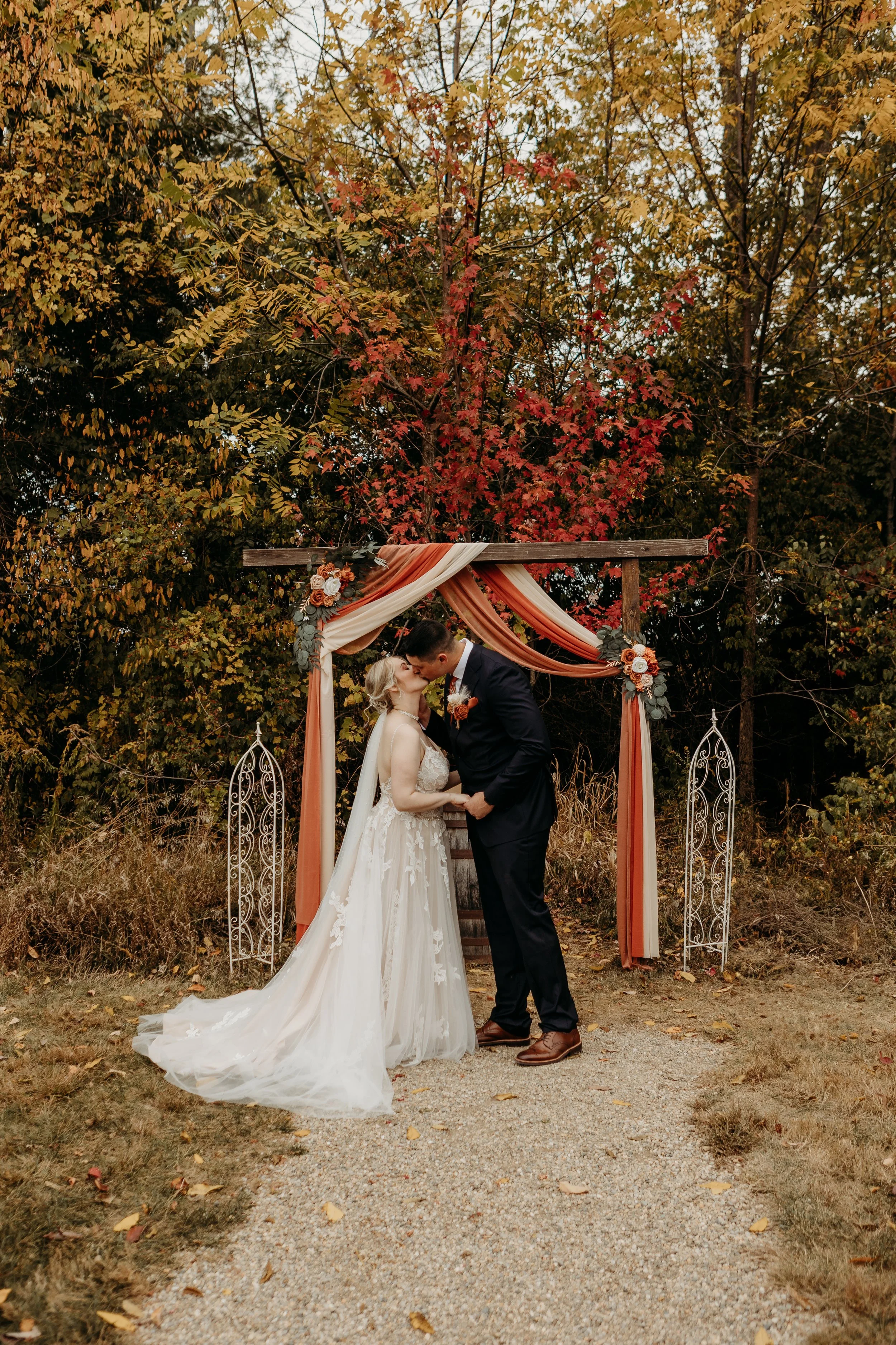 Wedding couple kissing under an autumn-themed altar with foliage and fabric drapes, outdoors in fall.
