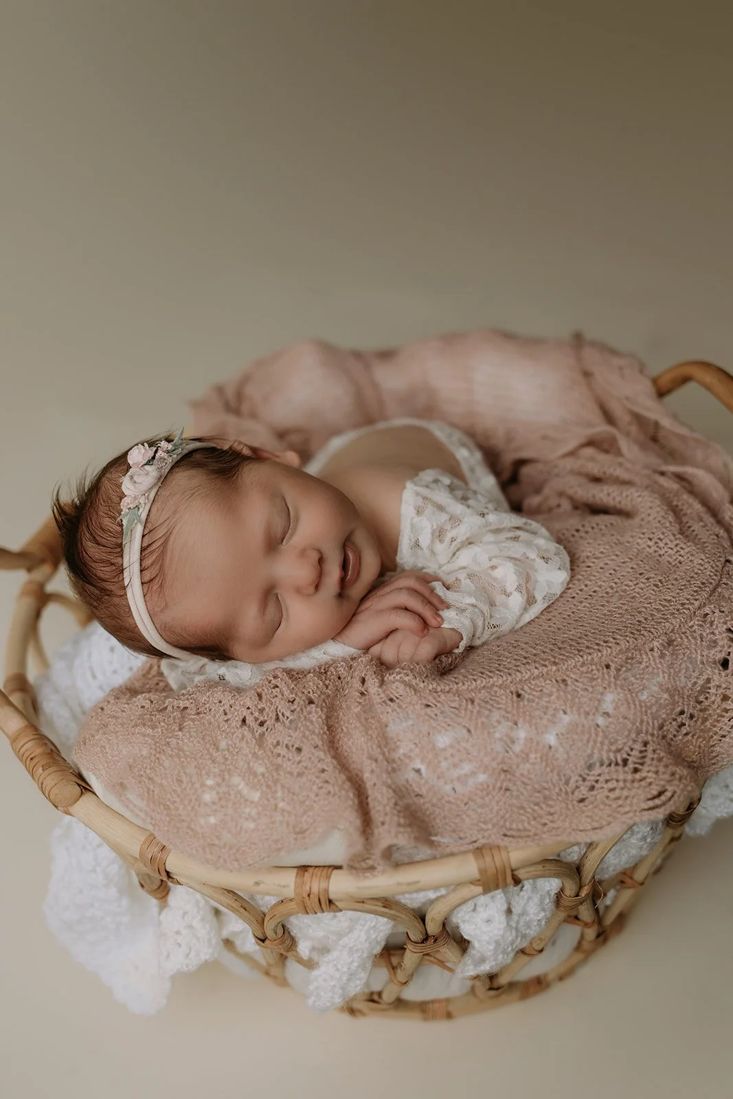 A sleeping baby with a floral headband, lying on a pink knit blanket inside a wicker basket.