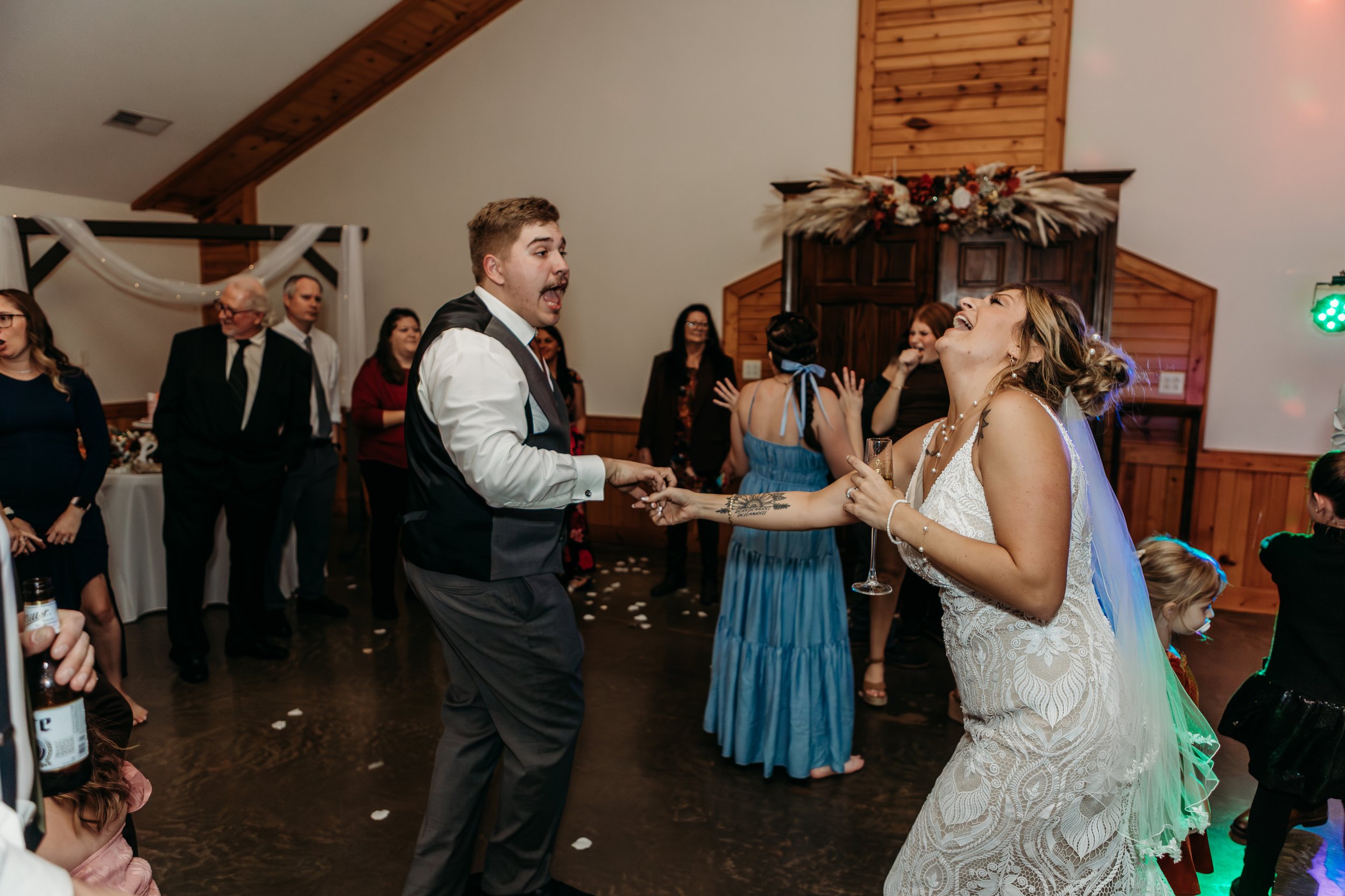 A bride and groom dancing and laughing at their wedding reception, with guests watching in the background.