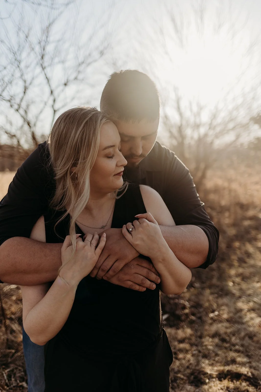 A couple embraces outdoors during sunset, with bare trees in the background, sharing a tender moment.