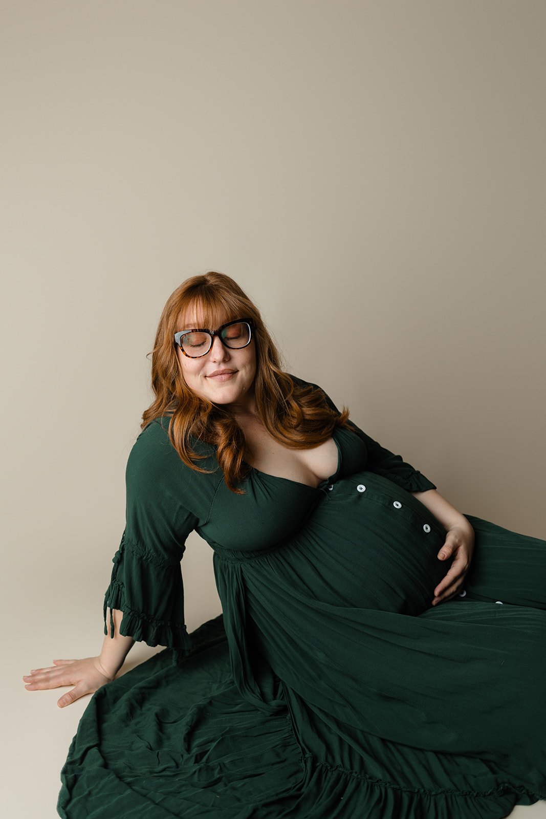 A pregnant woman with red hair, glasses, and a green dress is sitting on the floor with her eyes closed, smiling gently against a plain beige background.